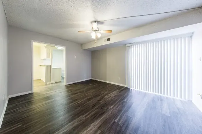 The Colony - living room with grey laminate wood flooring  with ceiling fan and sliding door with blinds to patio.