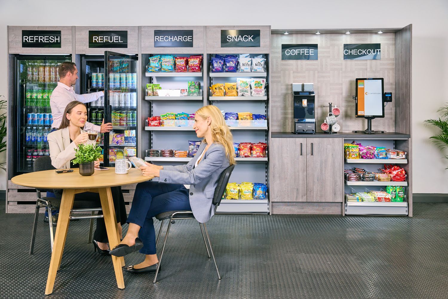 People at office: two women sit at table, man at fridge. Vending machines, coffee station, snacks.