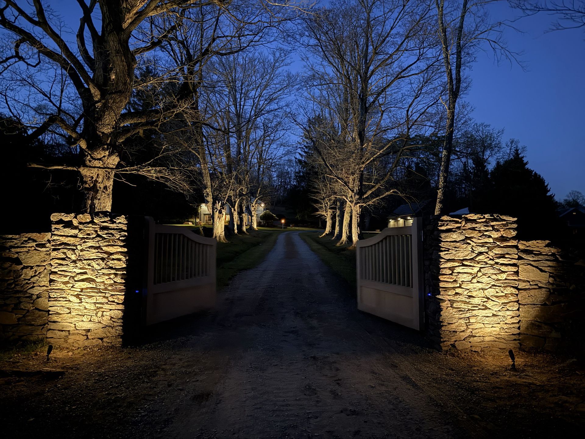 Stone gate lit at dusk opens to a long driveway lined with trees.