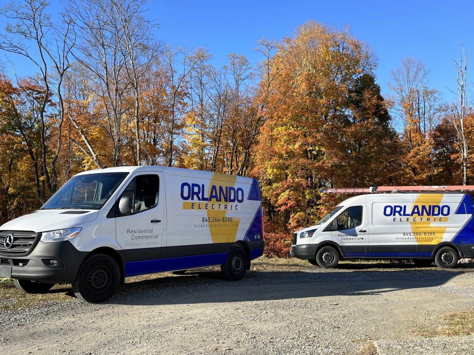 Two white Orlando Electric vans parked on gravel road with fall foliage.