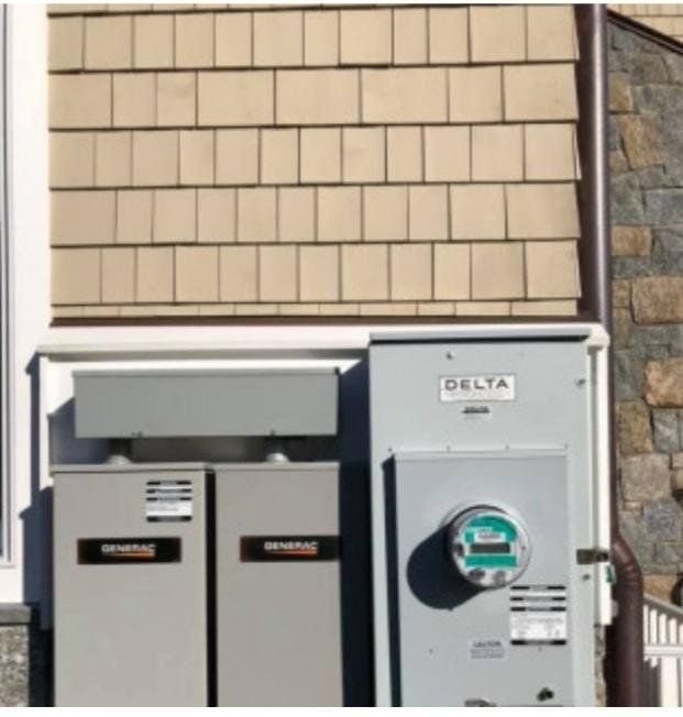 Two gray electrical boxes and a meter are mounted on a house exterior with tan shingles.