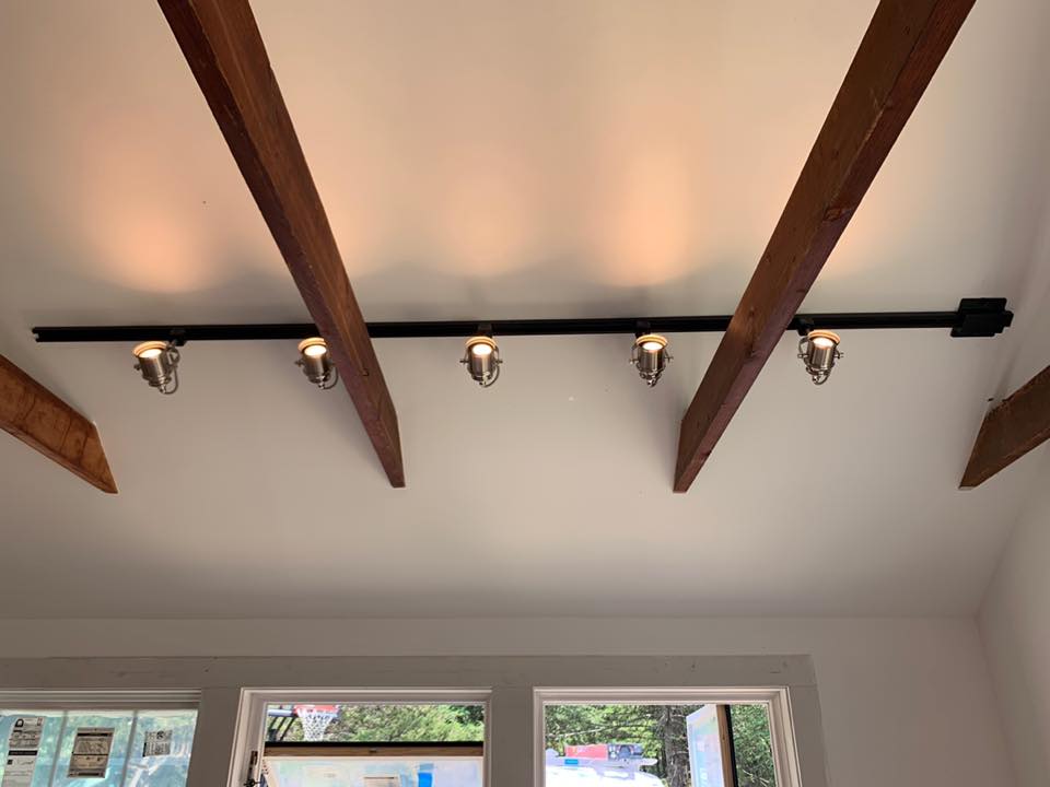 Black track lighting on a white ceiling with wooden beams, illuminating the space below.