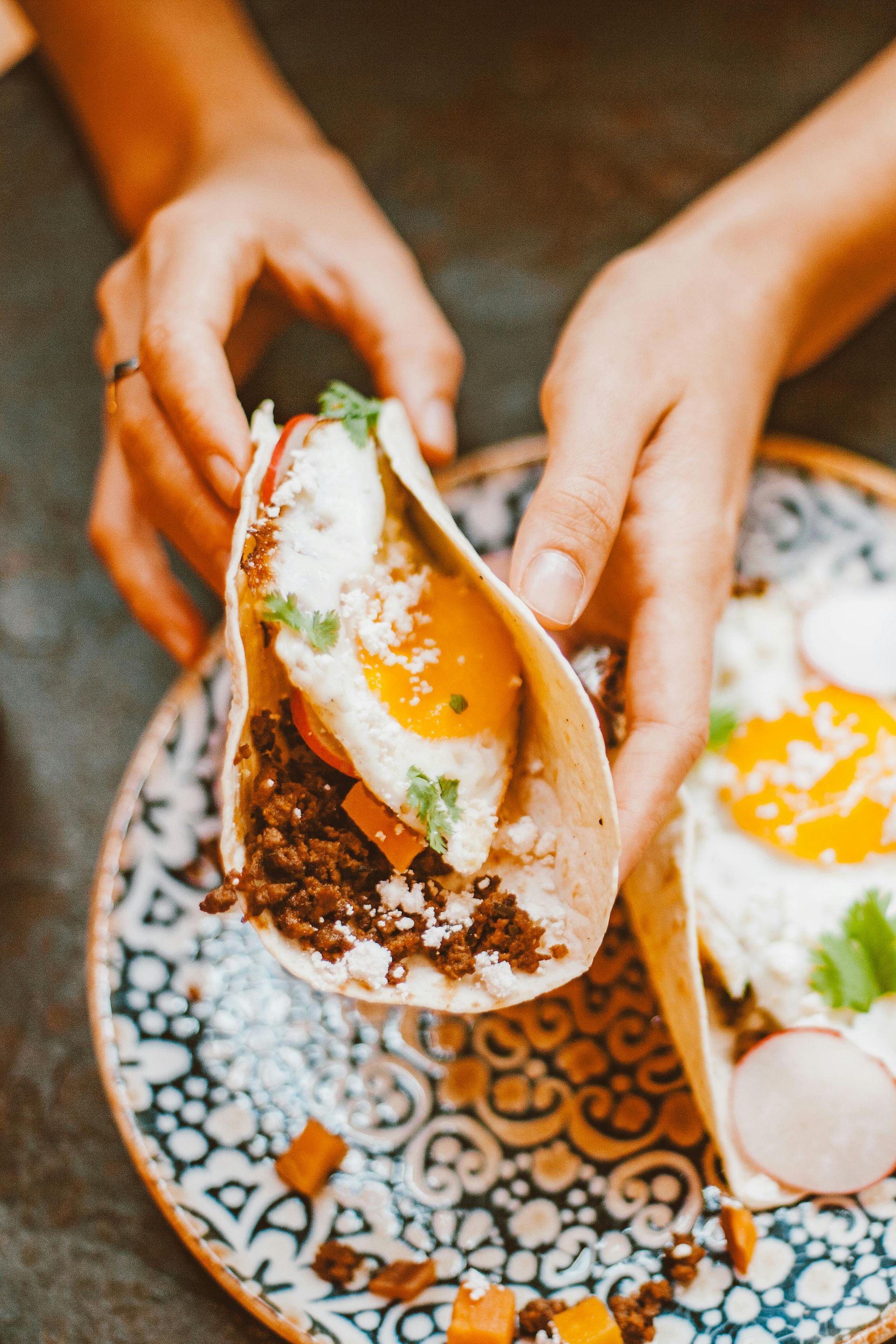 Person holding a taco with an egg, meat, and cheese on a patterned plate.