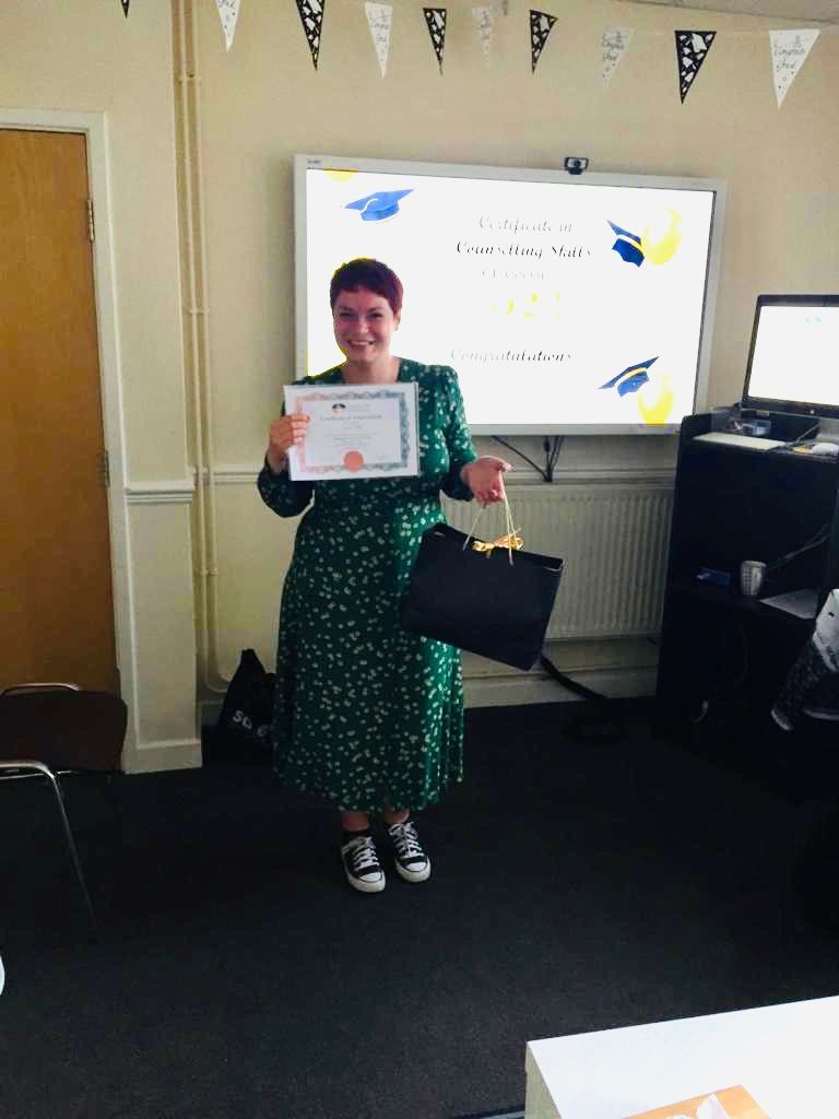 Woman in green dress holds certificate, smiling in a classroom.