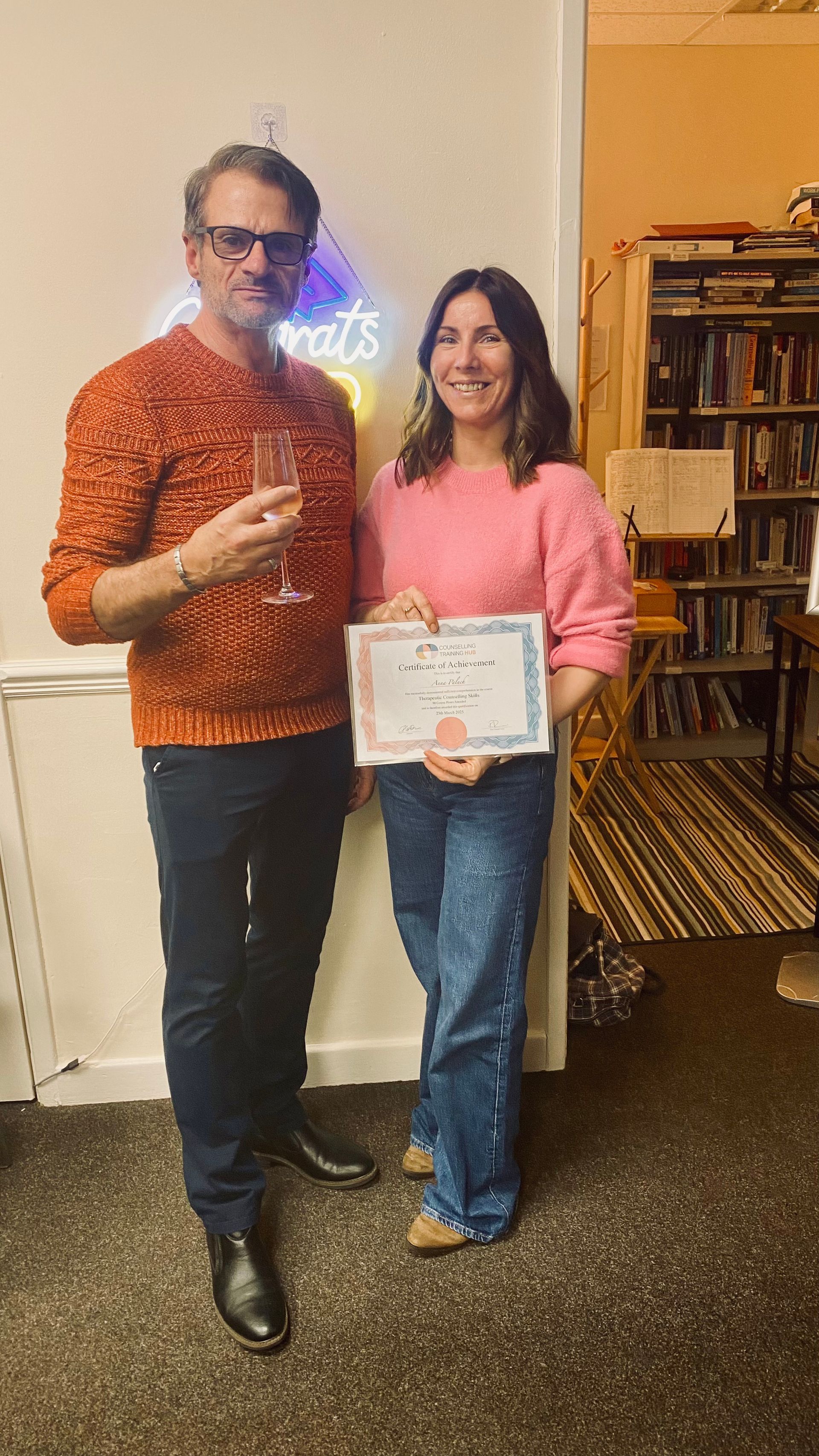 Man in orange sweater and woman in pink sweater holding a certificate in a room with a 