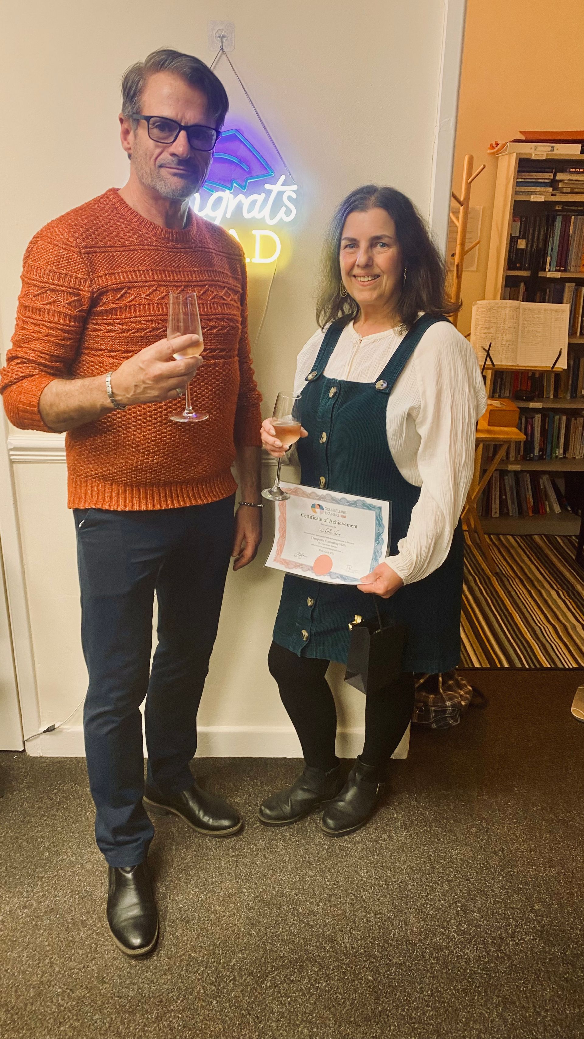 Man and woman holding champagne, woman holding certificate. Neon sign in background.