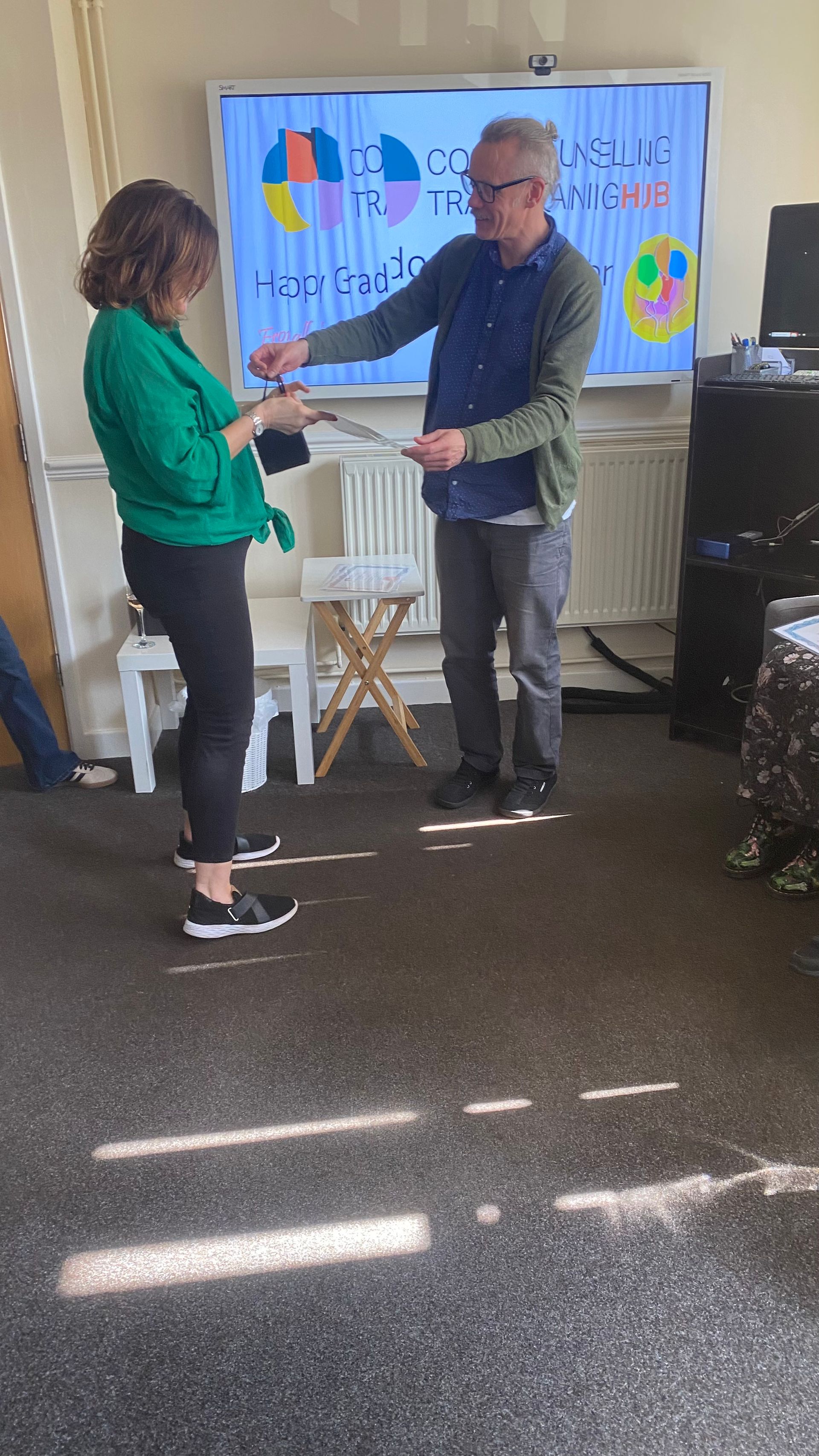 Two people at a presentation. Man in gray pants and a blue shirt, holding papers. Woman in green top, holding papers. Whiteboard behind them.