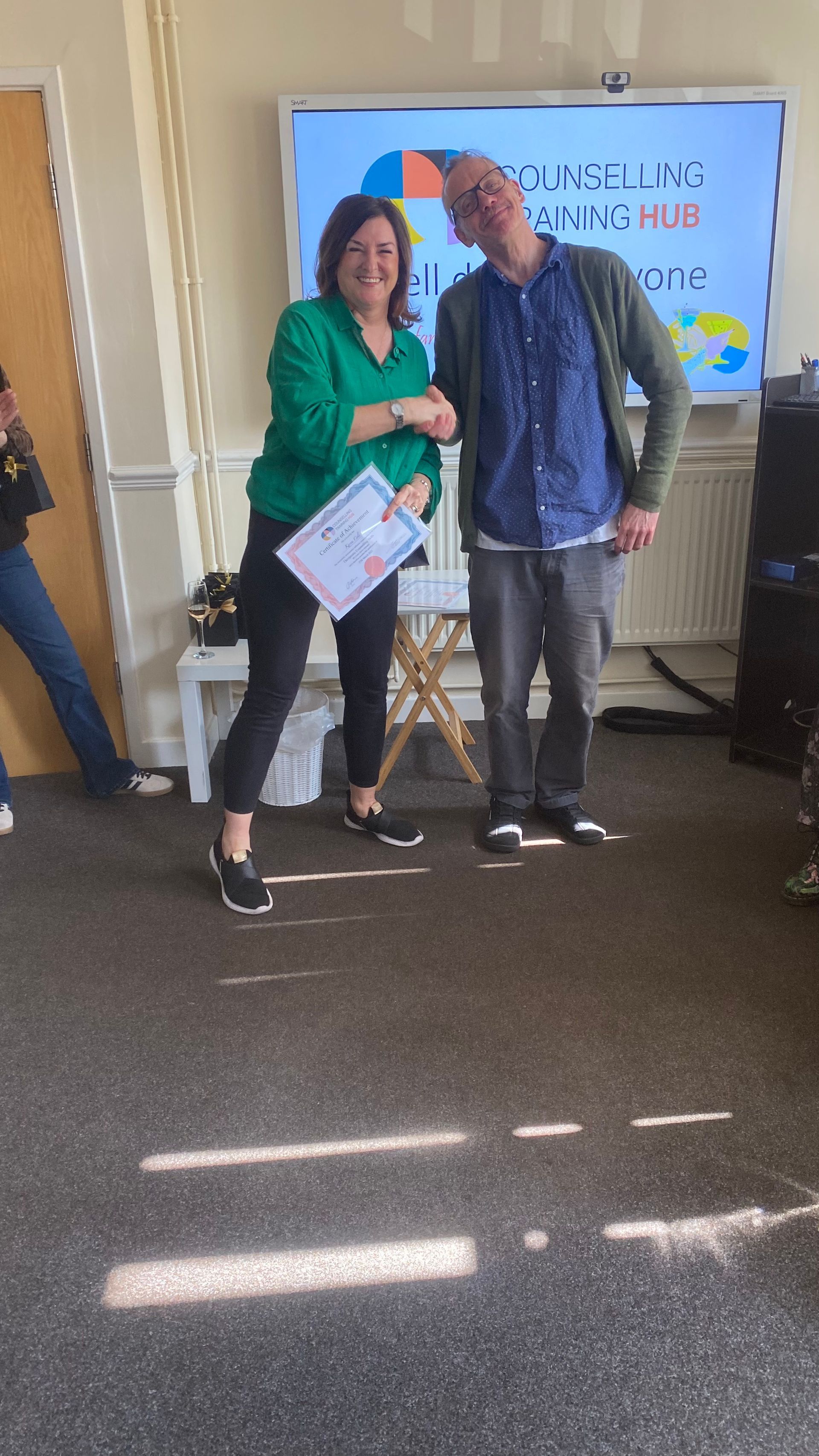 Woman in green shirt handing a certificate to a man in blue shirt, in a room with a presentation screen.