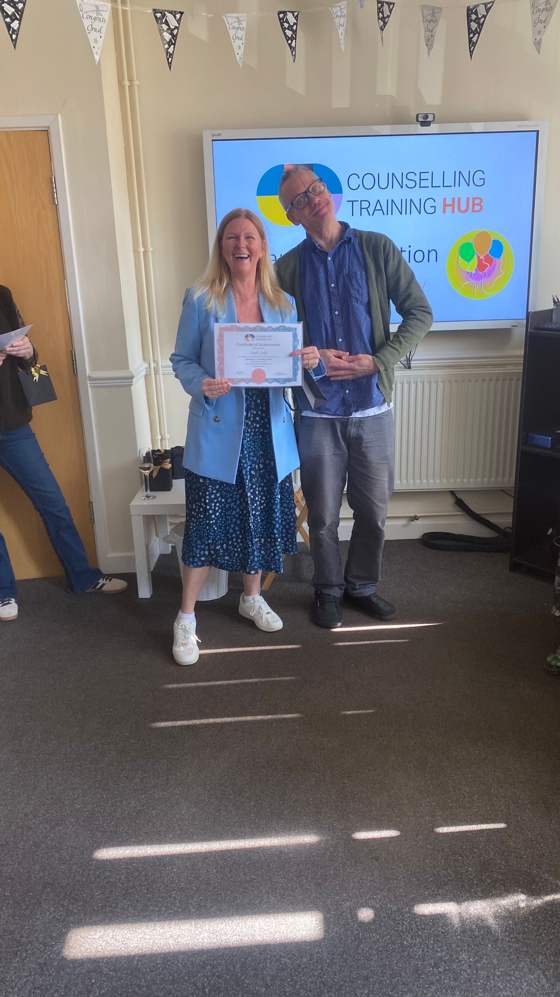 Woman in blue dress and man in blue shirt standing, smiling, with a certificate, in front of a screen.