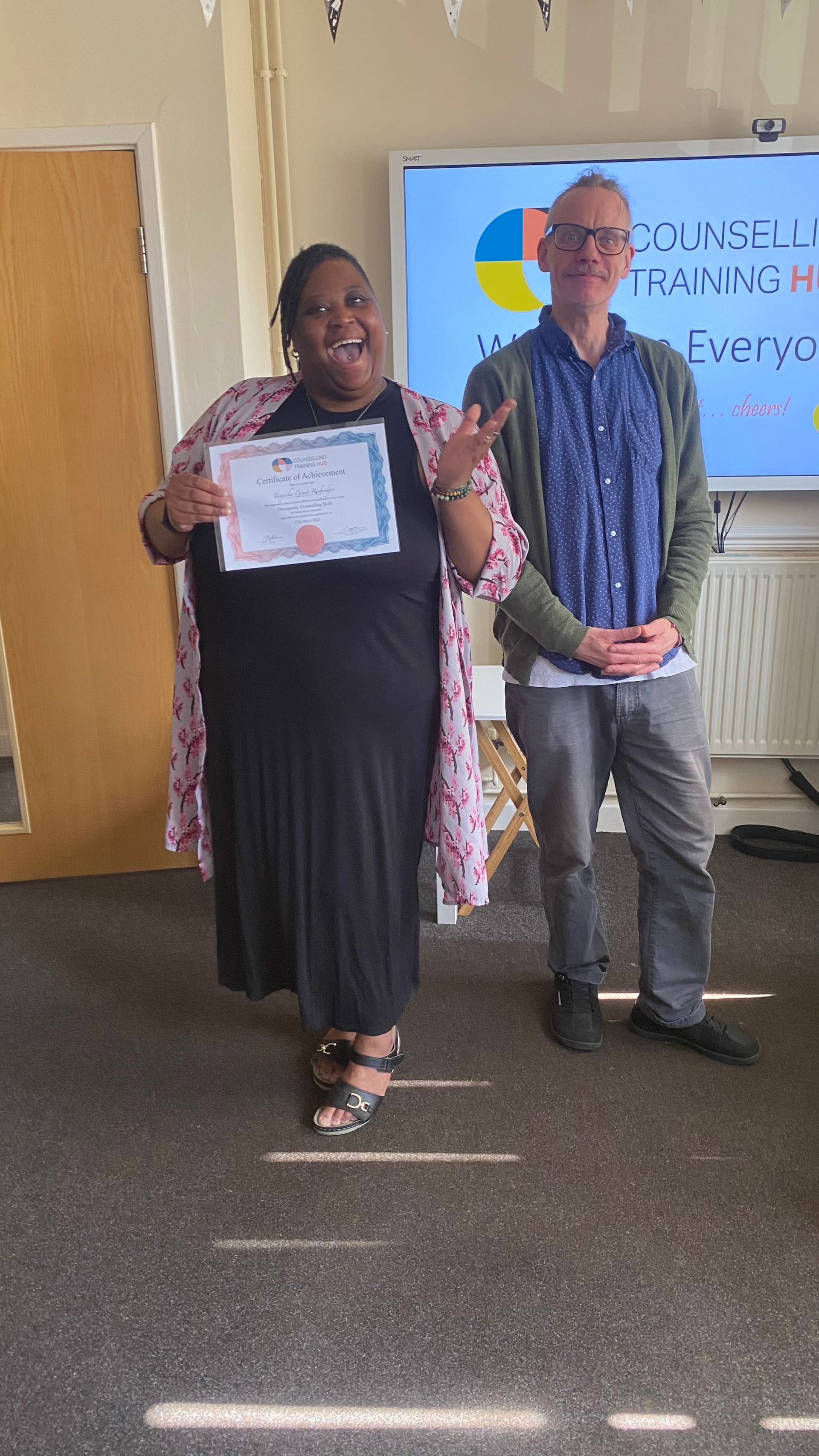 Woman holding certificate smiles, man stands beside her, in front of a whiteboard.