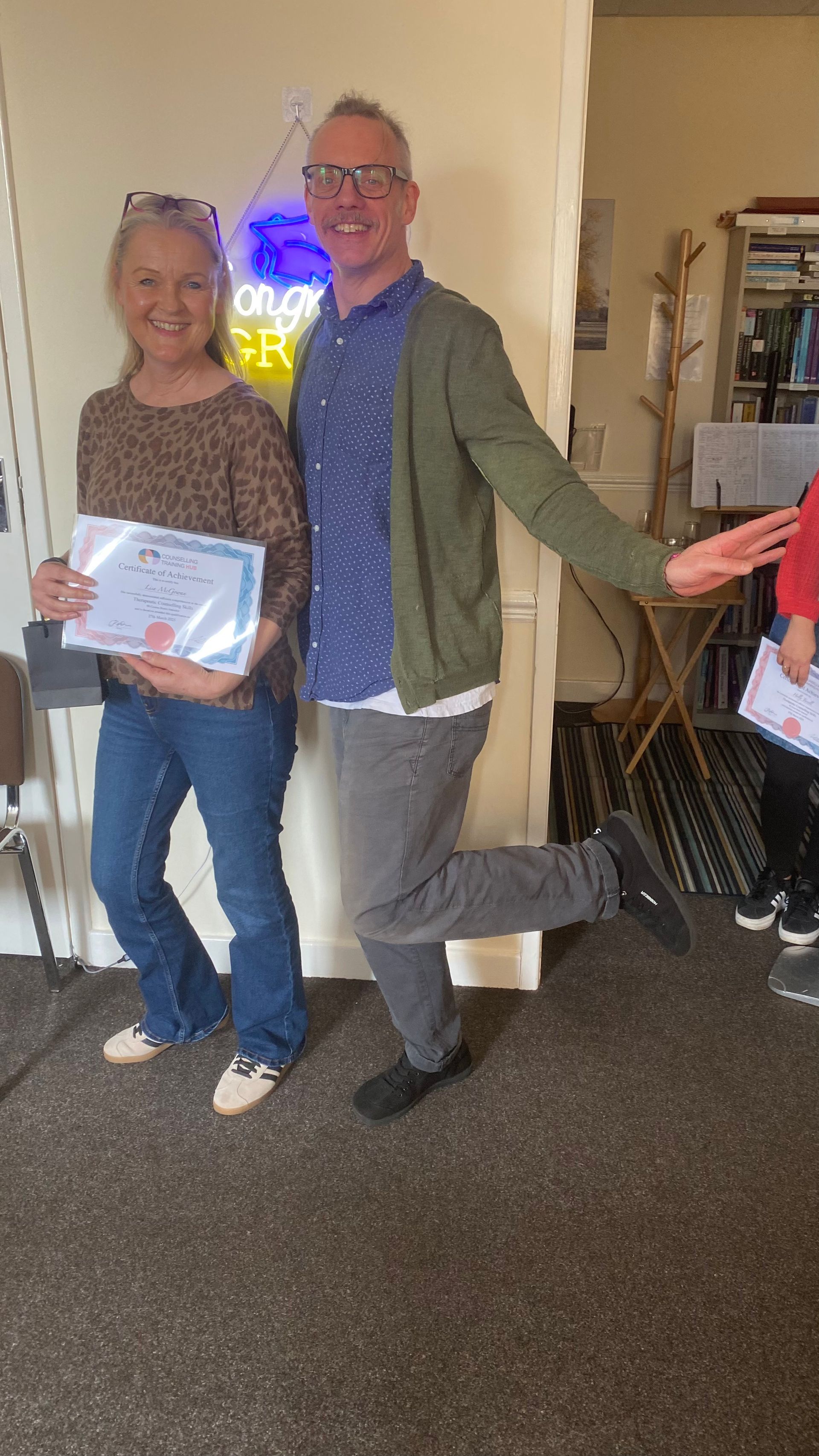 Woman and man holding certificates, posing. Man has leg raised, both smiling near a neon sign.