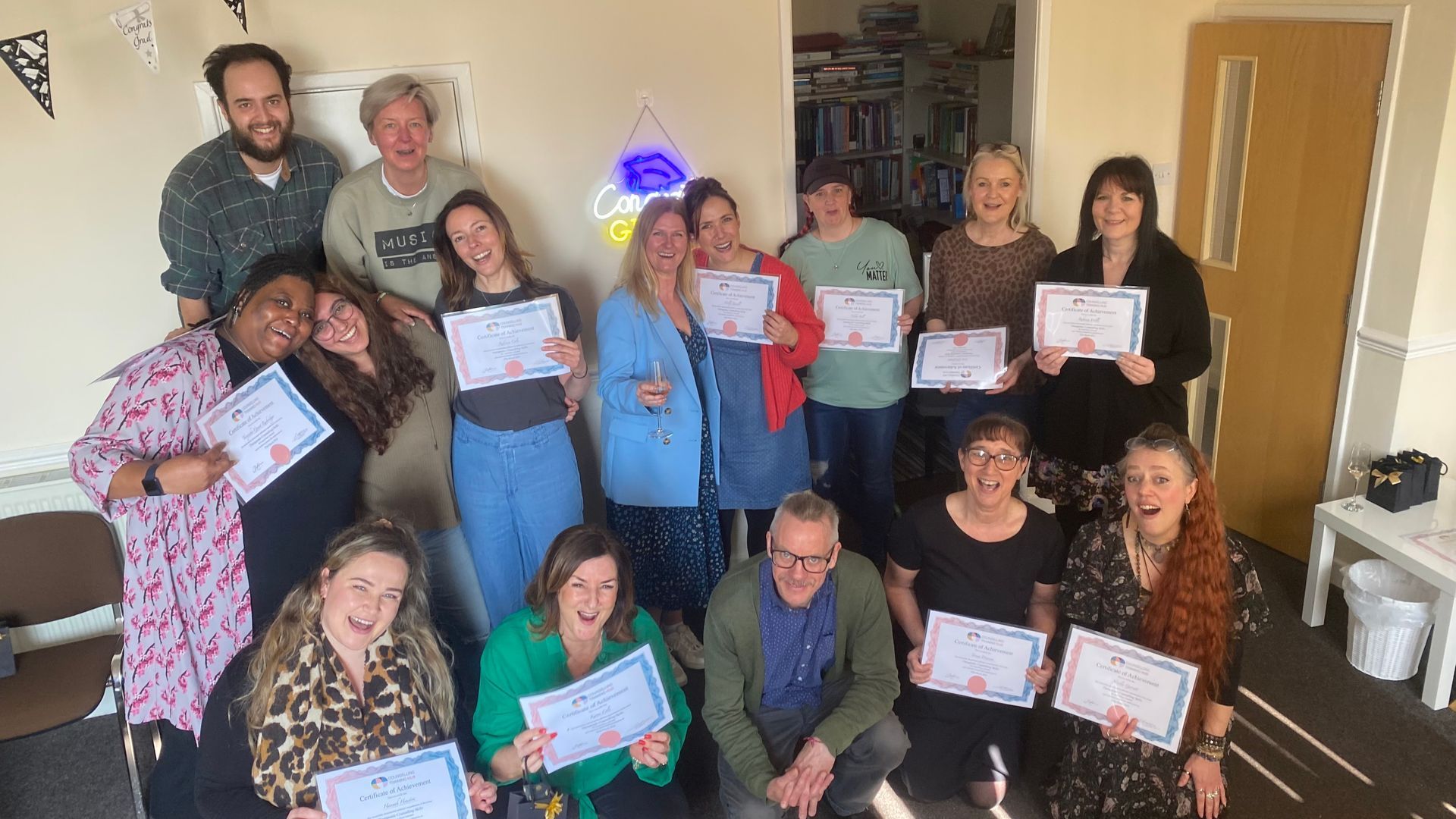 Group of people holding certificates, smiling, in a room. Some are embracing. A blue and yellow light is in the background.