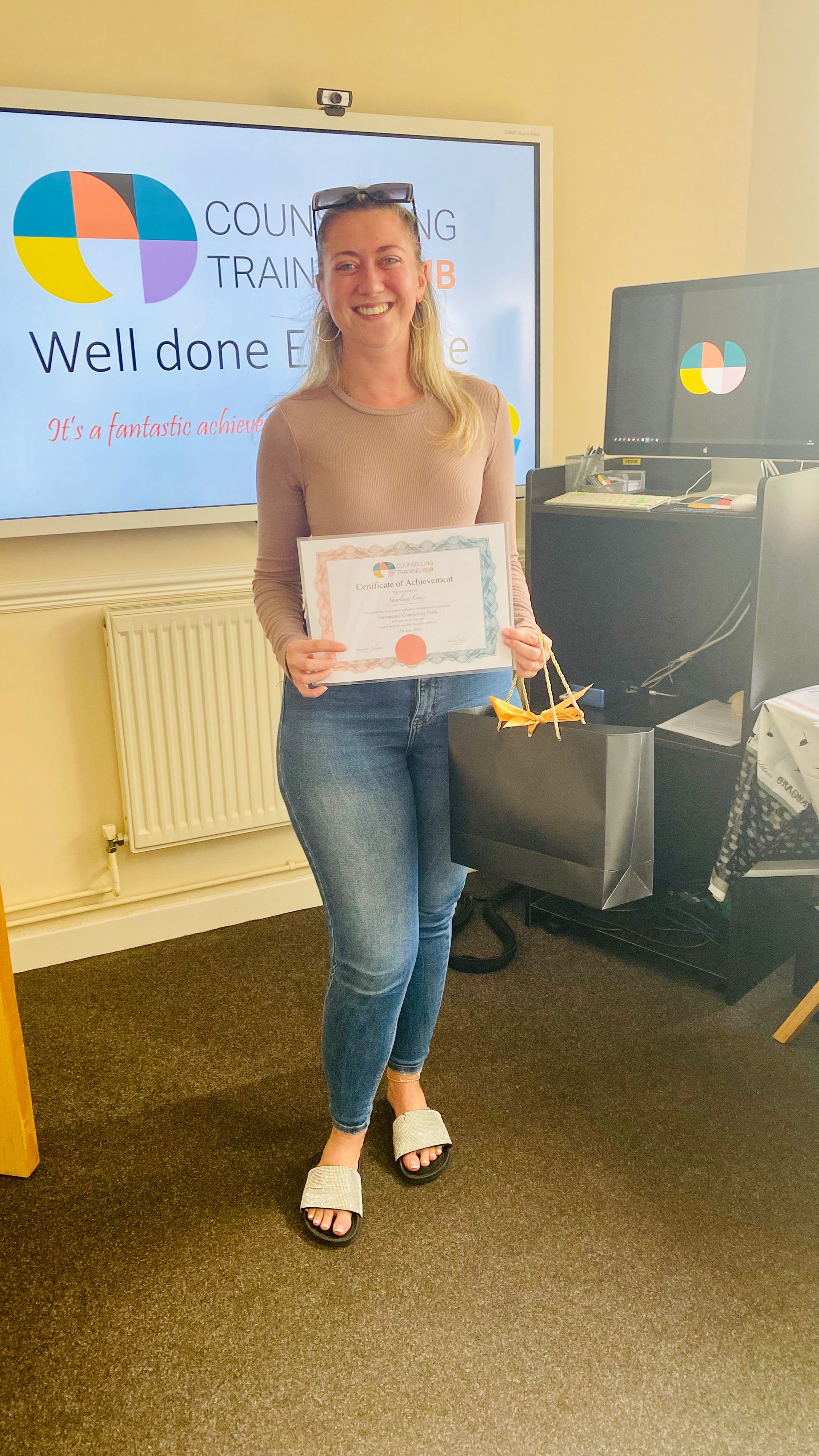 Woman smiling, holding certificate and gift bag. Standing in front of a screen with a logo and the words 