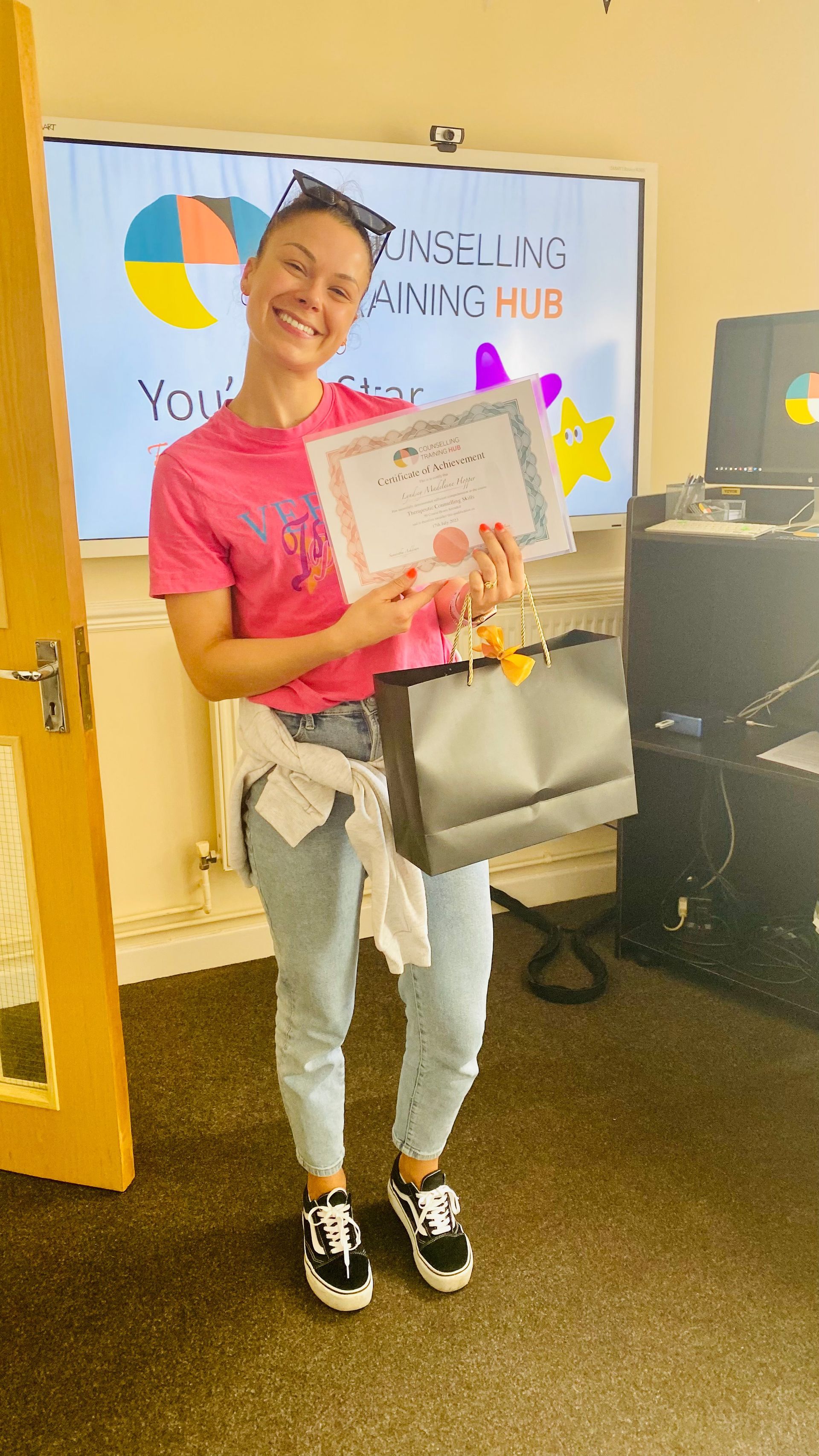 Woman holding a certificate and gift bag, smiling. Standing in front of a training hub sign.