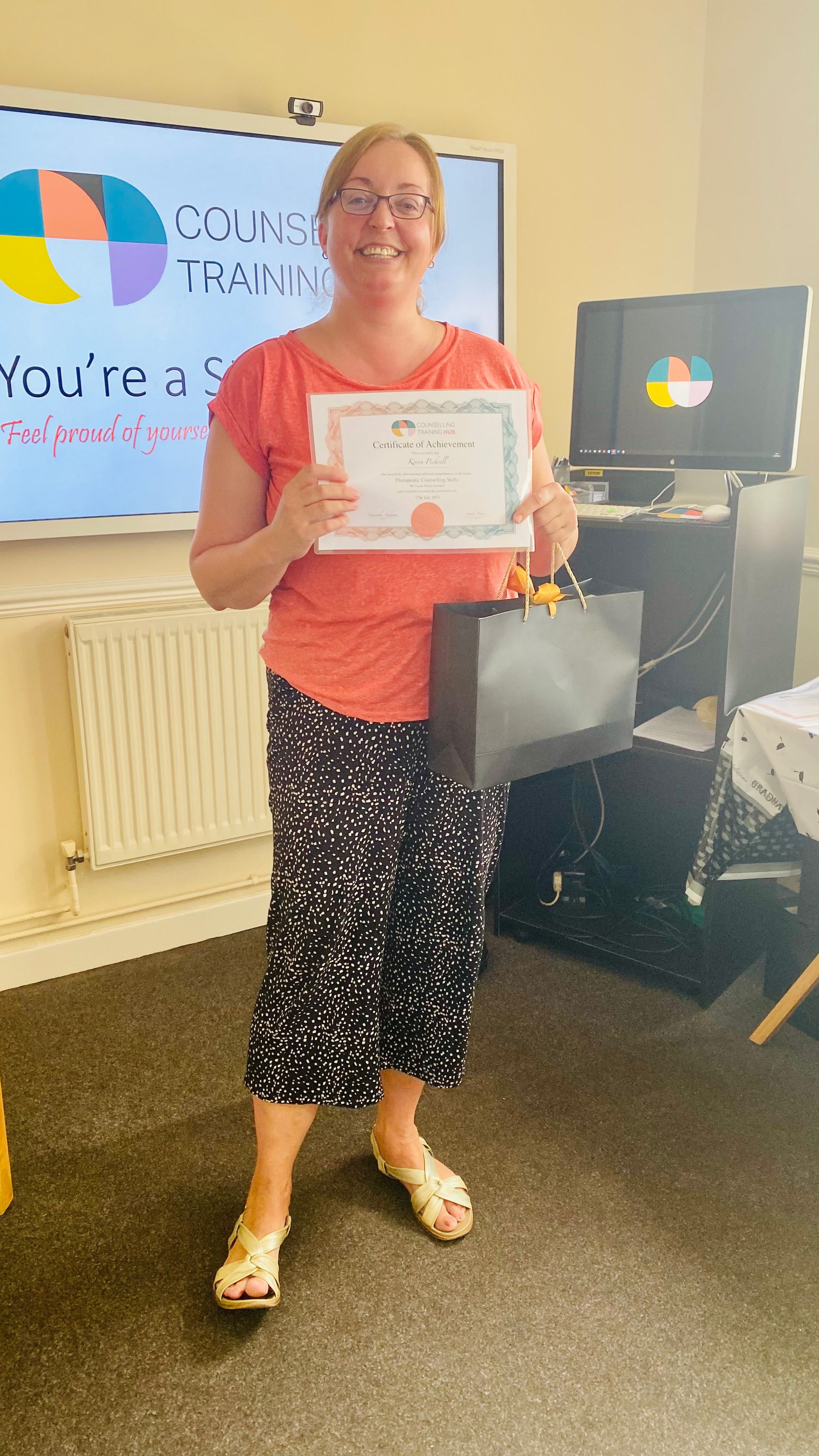 Woman in coral shirt and patterned pants holding a certificate and gift bag, in front of a whiteboard.