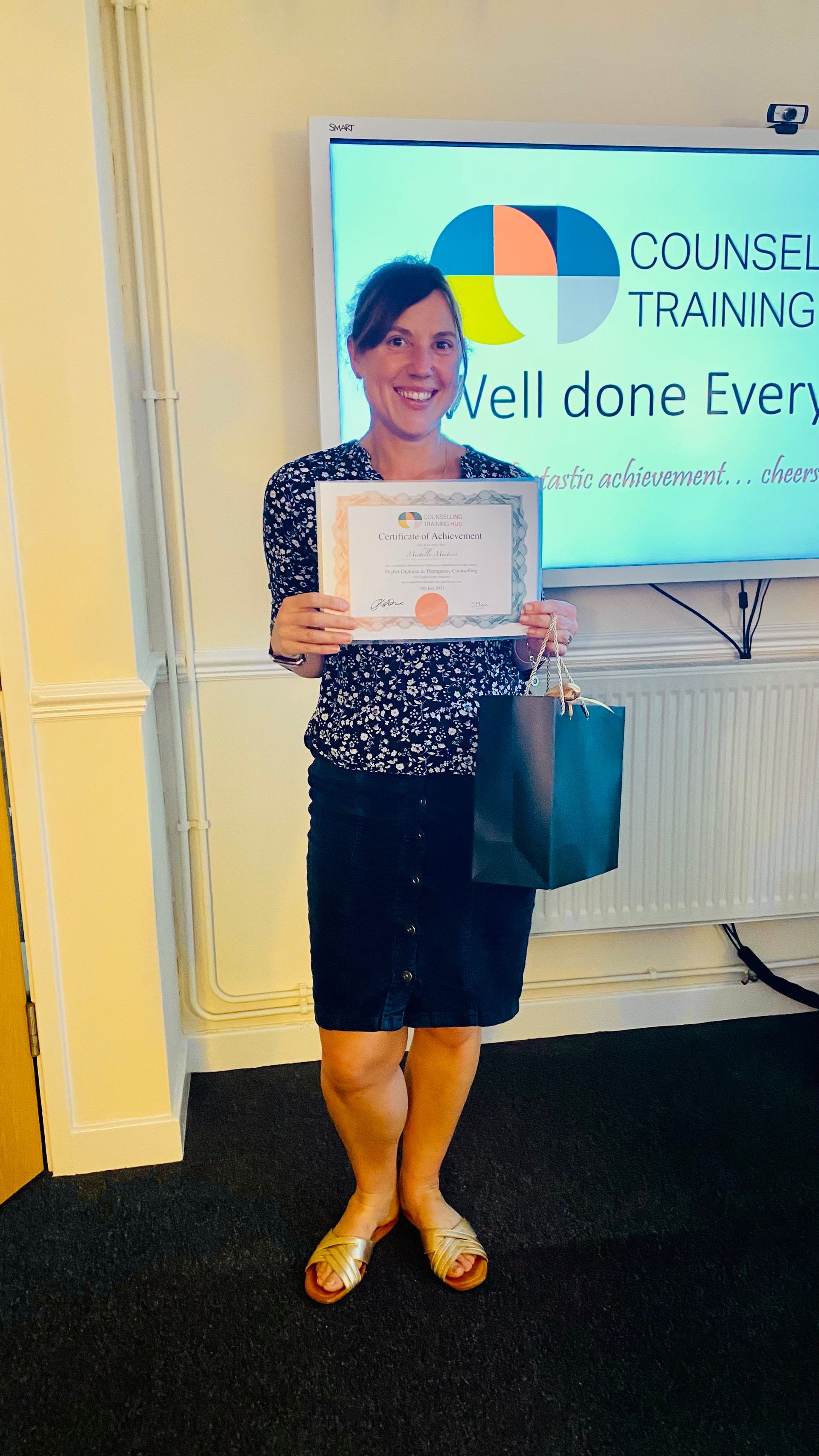 Woman holding a certificate and gift bag, smiling in front of a whiteboard that says 
