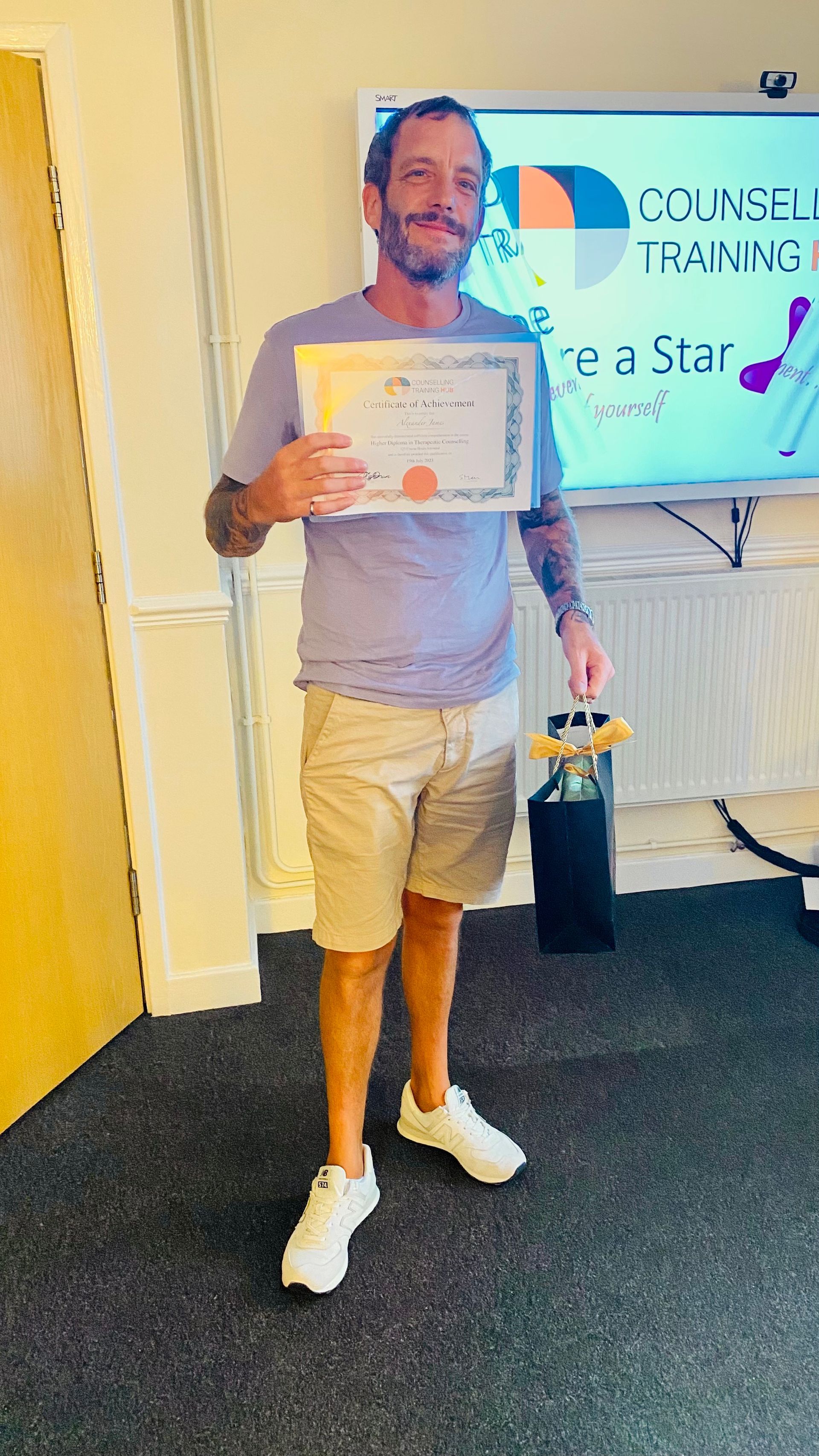 Man holding a certificate and gift bag, standing in a room with a presentation screen.
