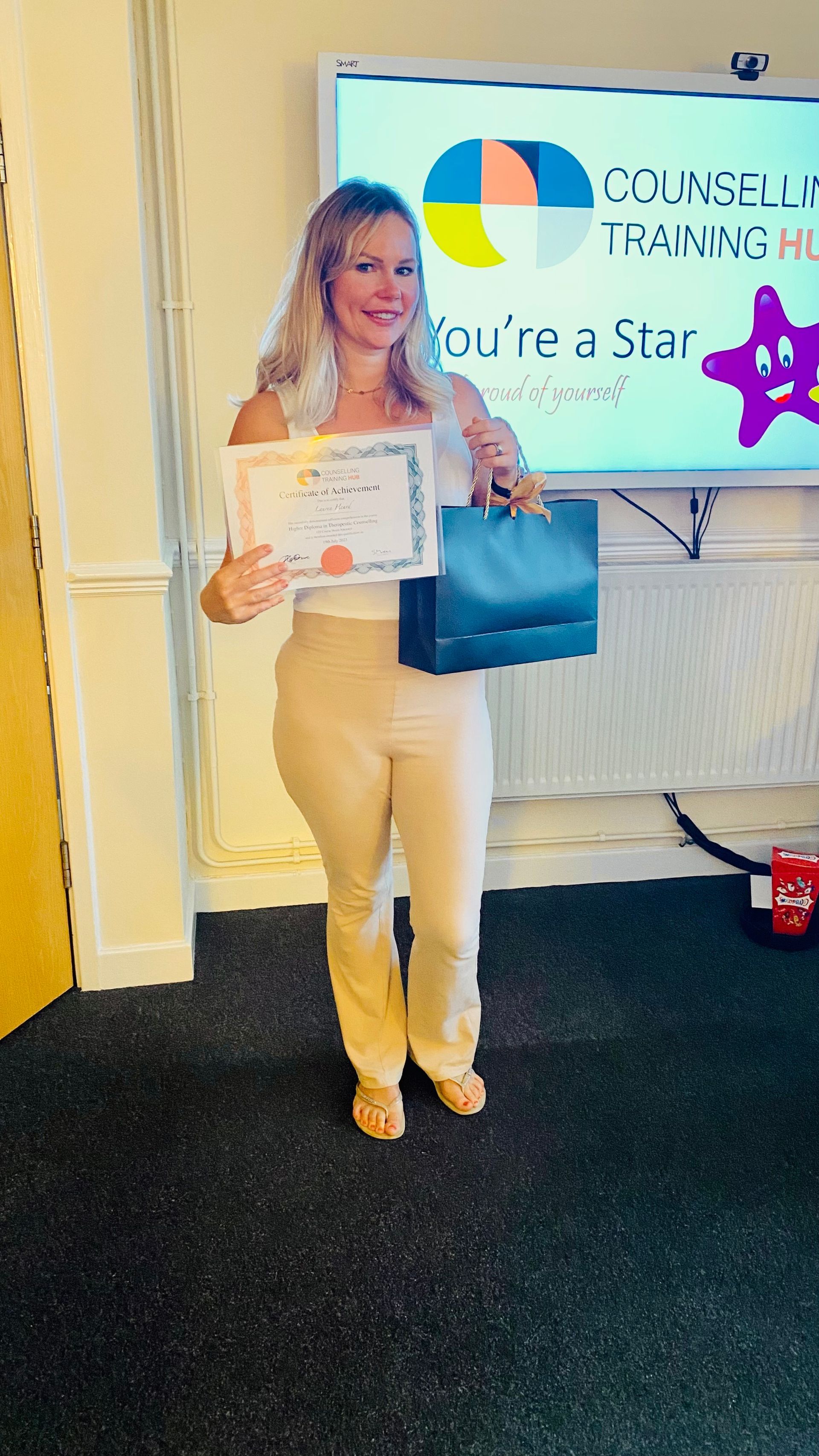 Woman holding a certificate and gift bag, standing in front of a screen with a 