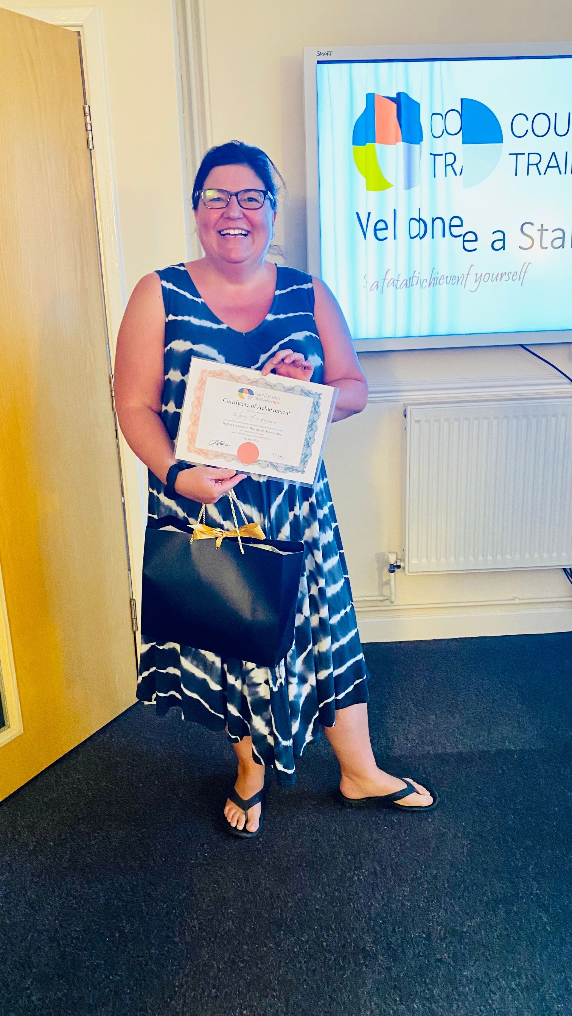 Woman in a blue and white dress holding a certificate, smiling. Standing in front of a sign.