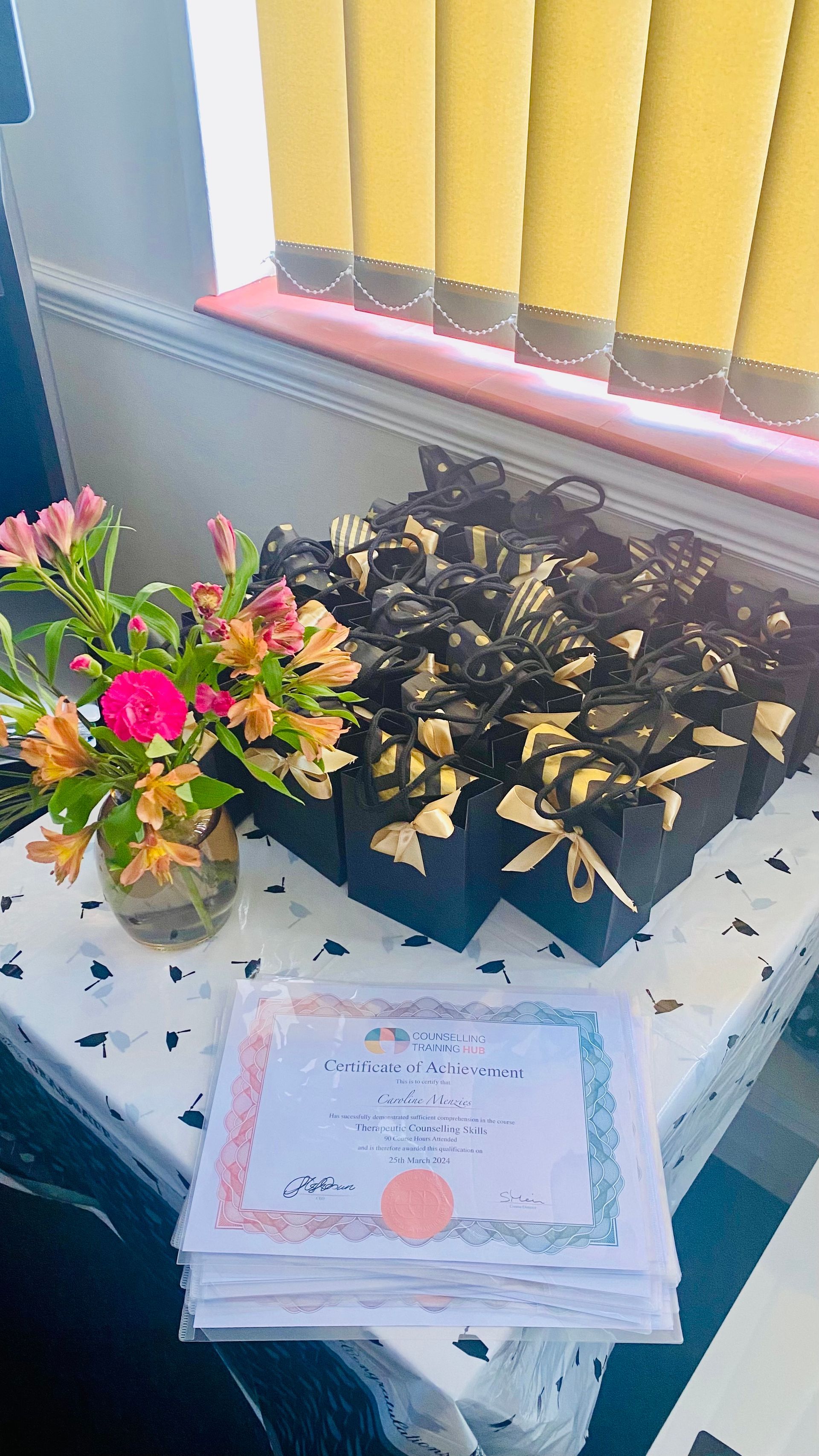 Certificates and black rose boxes on a table, next to a flower arrangement by a window.