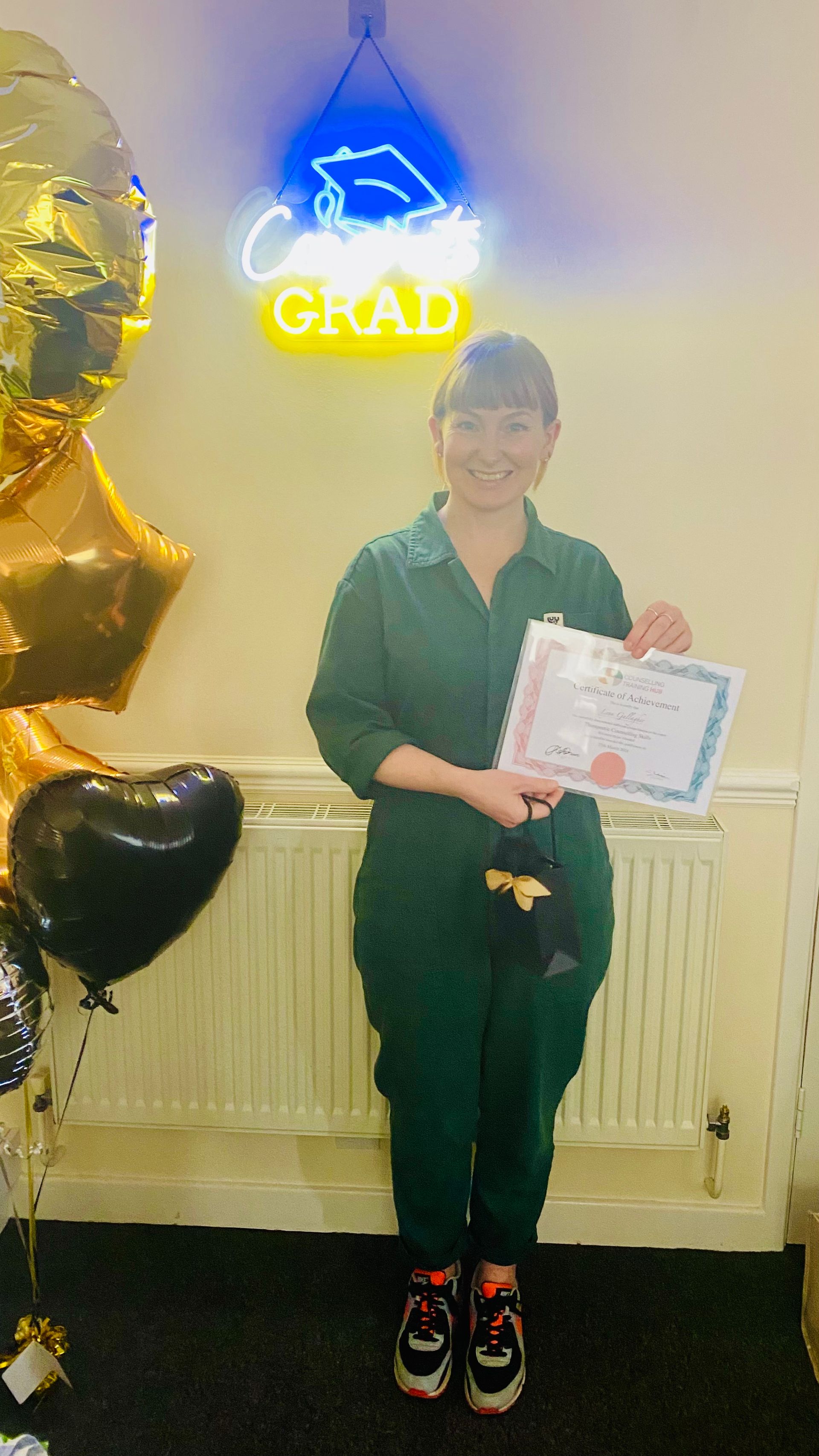 Woman in green jumpsuit holding certificates, standing in front of balloons and a 