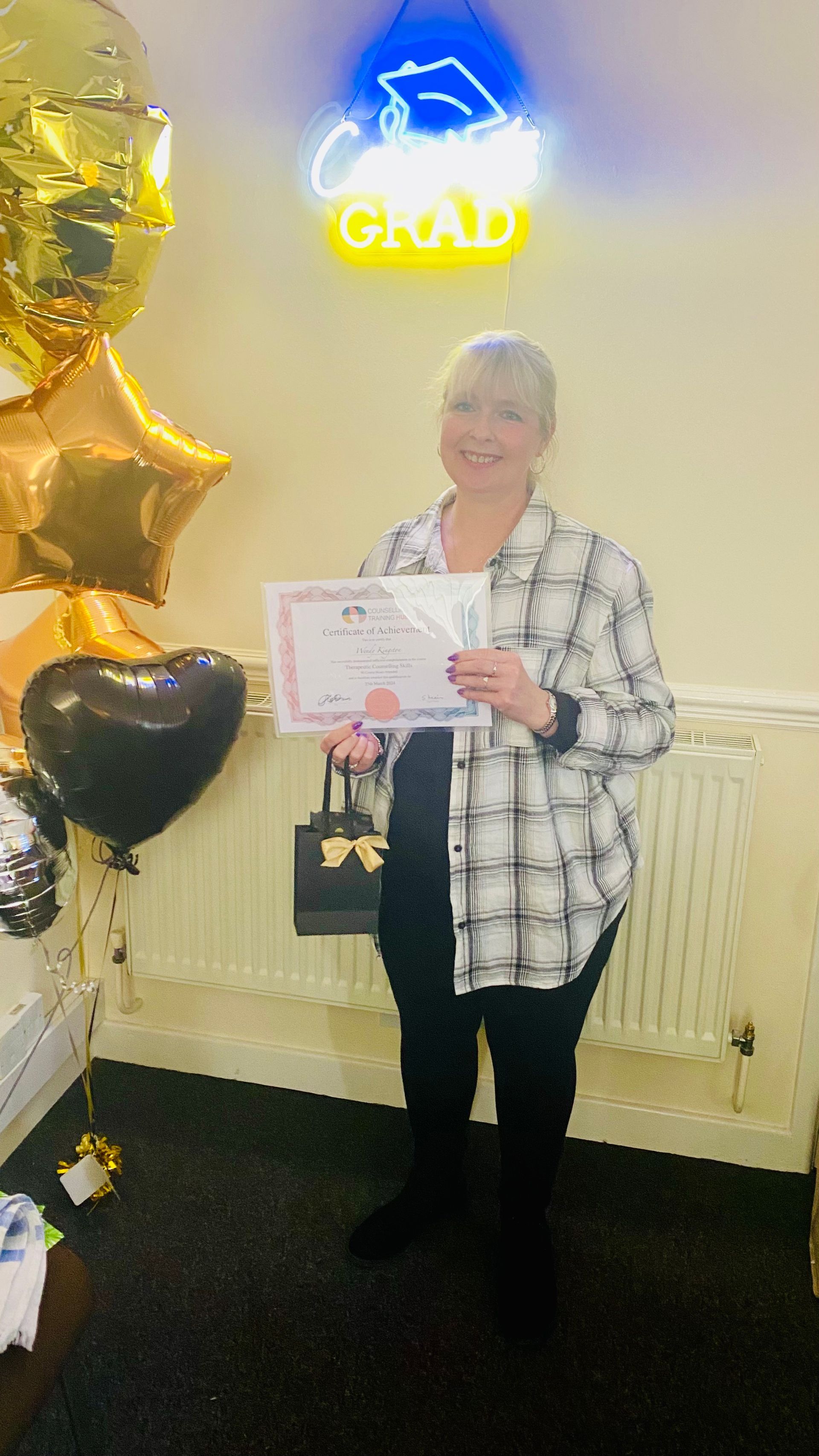 Woman holding a certificate at a graduation celebration, with balloons and neon sign that reads 