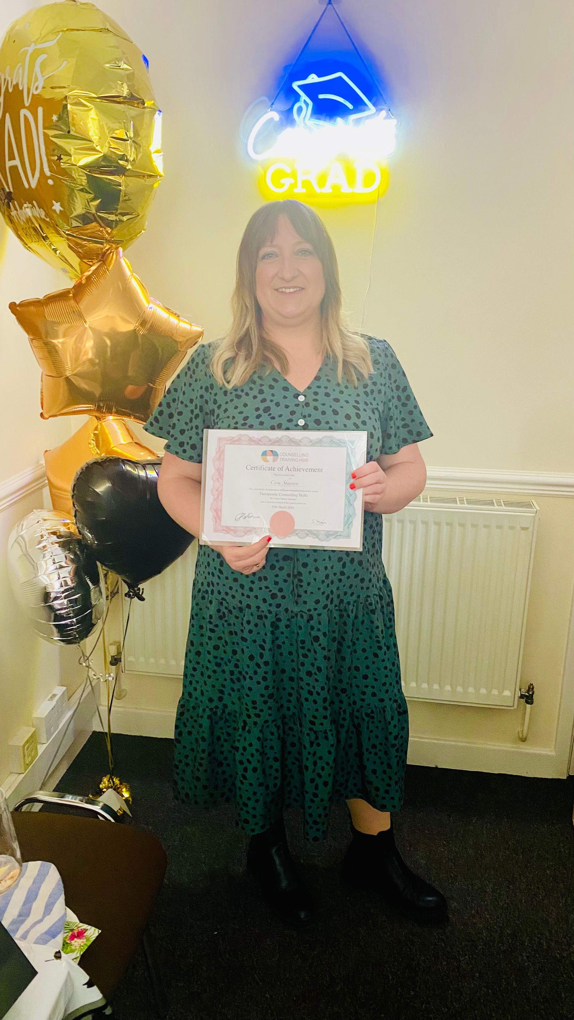 Woman in green dress holding a certificate, celebrating graduation with balloons and neon sign.