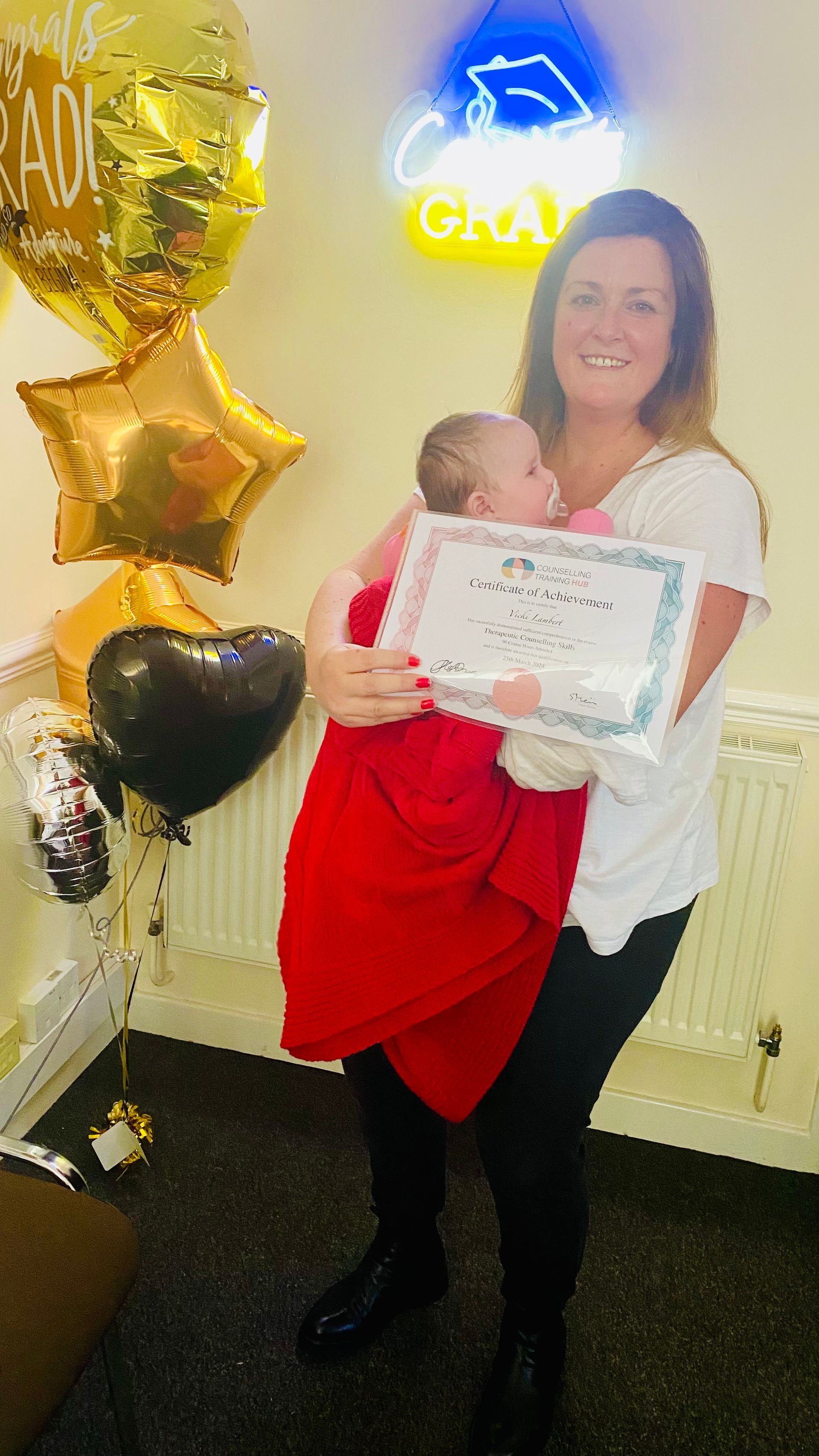 Woman holding baby wrapped in red blanket, showing certificate, with graduation balloons and neon sign in background.
