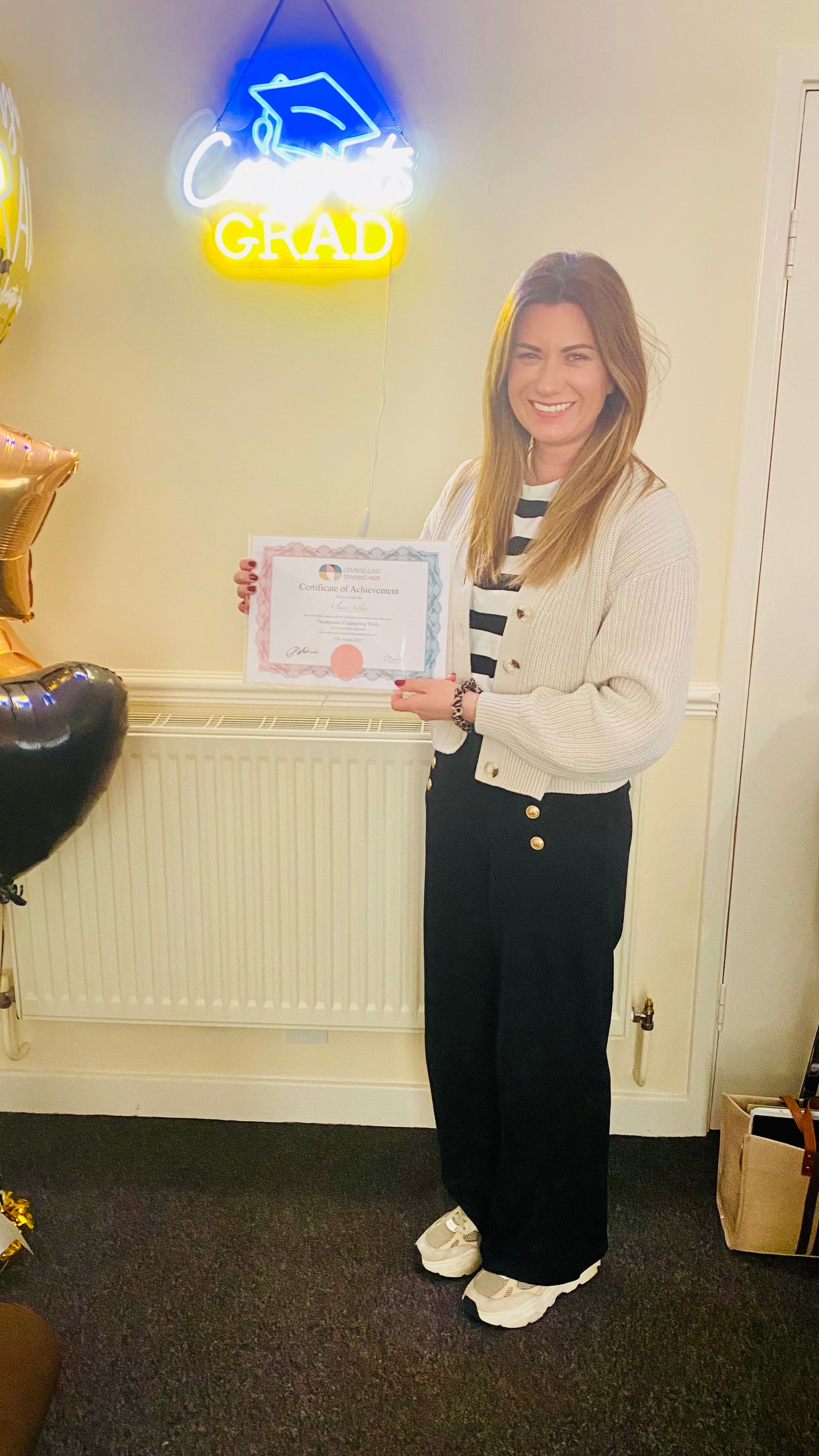 Woman in black pants and white cardigan holds a certificate in front of a graduation sign.