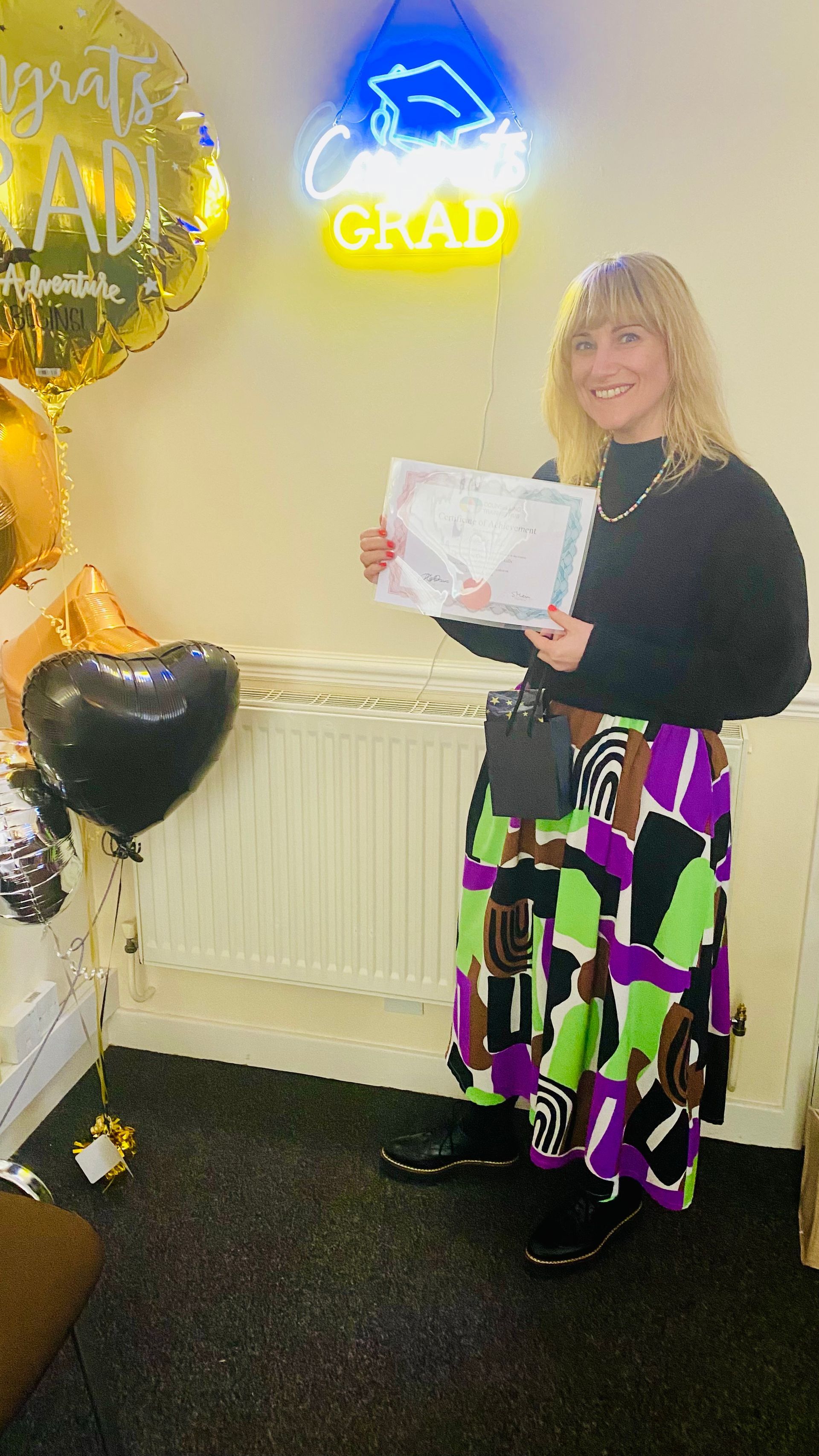 Woman holding a diploma, smiling, by balloons and a 