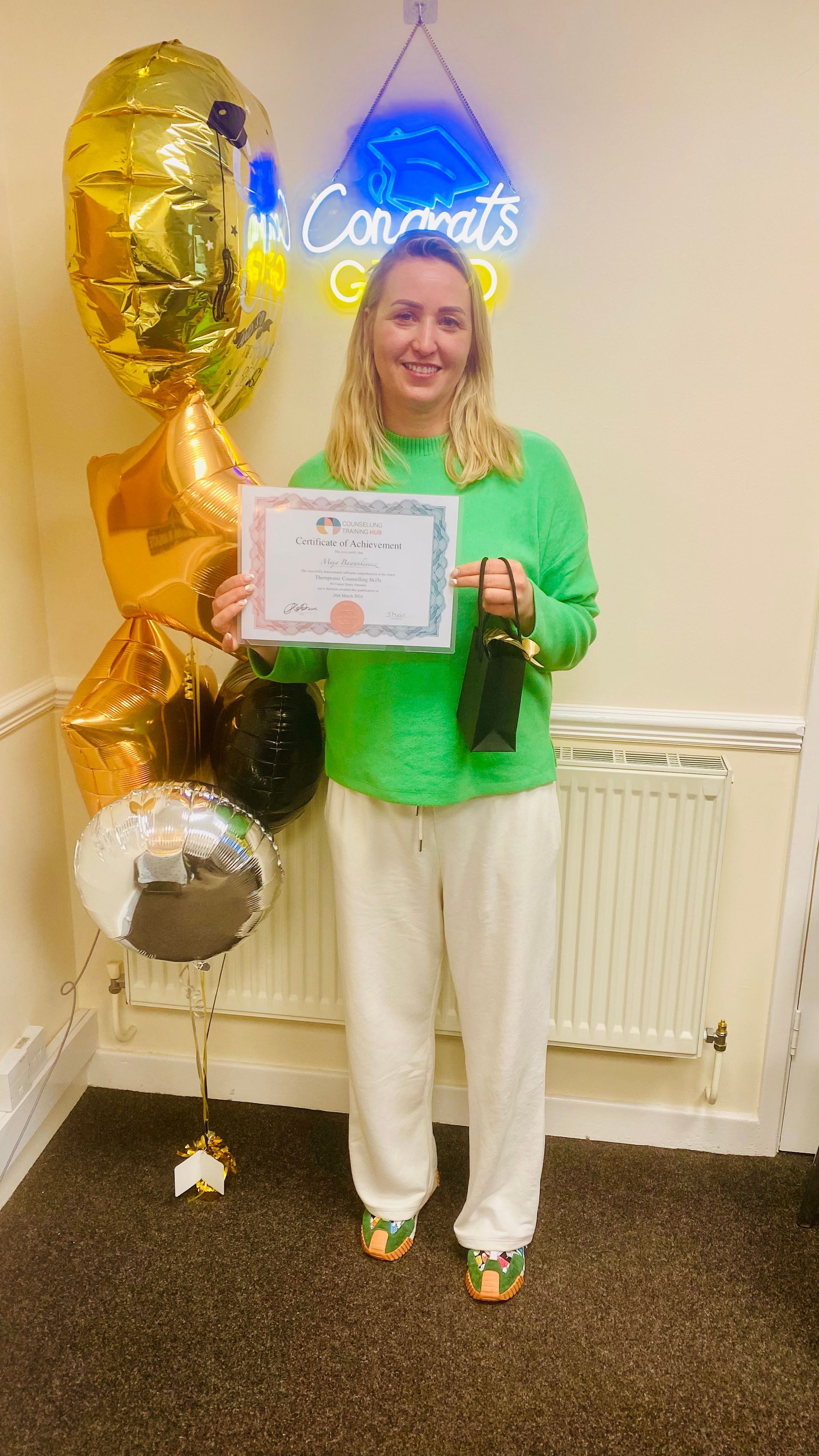 Woman in green sweater and white pants, holding a certificate. Balloons and 
