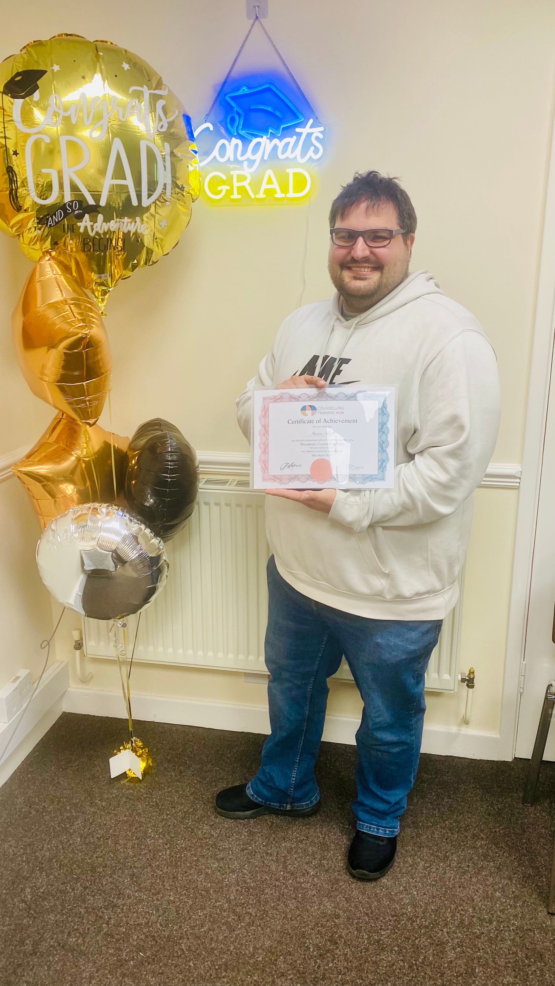 Man holding a certificate, smiling near graduation balloons and a 