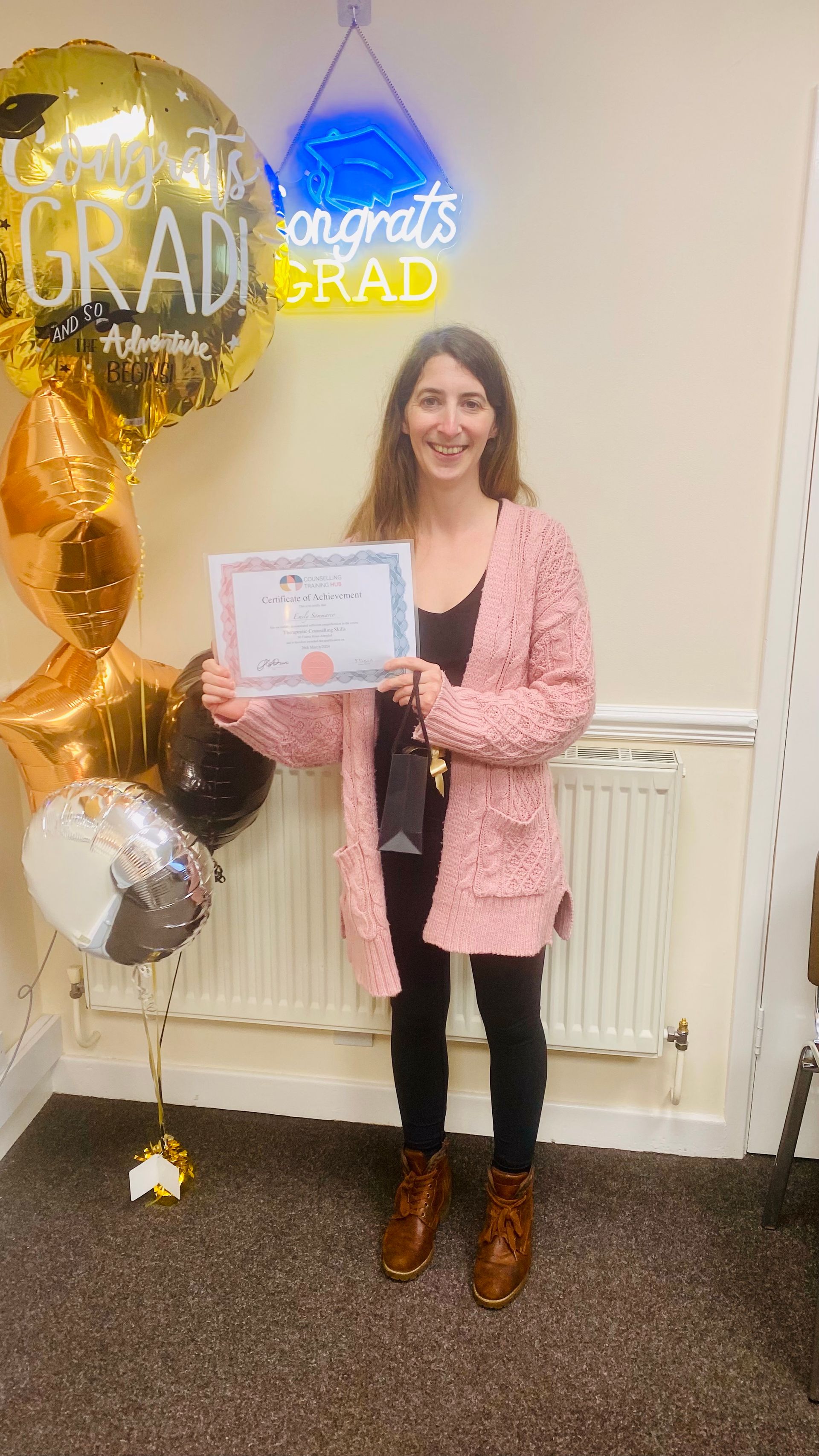 Woman in pink cardigan holds a certificate, standing by balloons and a 