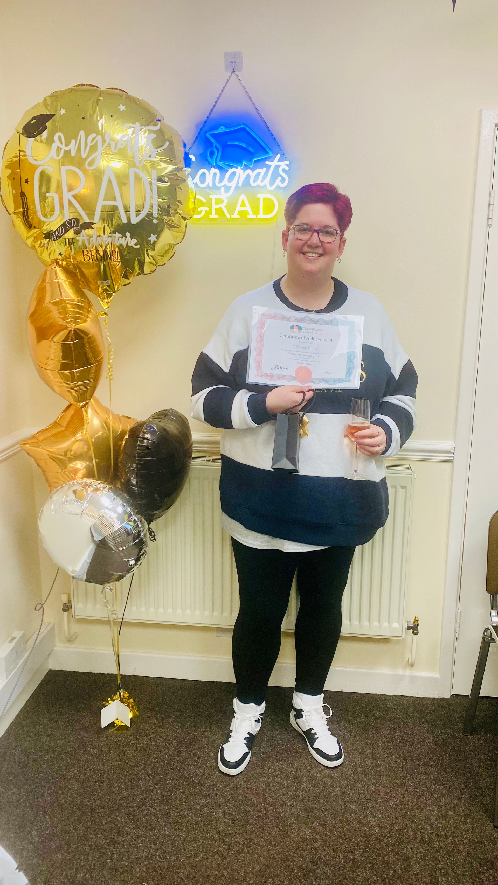 Woman in black pants and sweater holds diploma, trophy, and smiles. Balloons and 