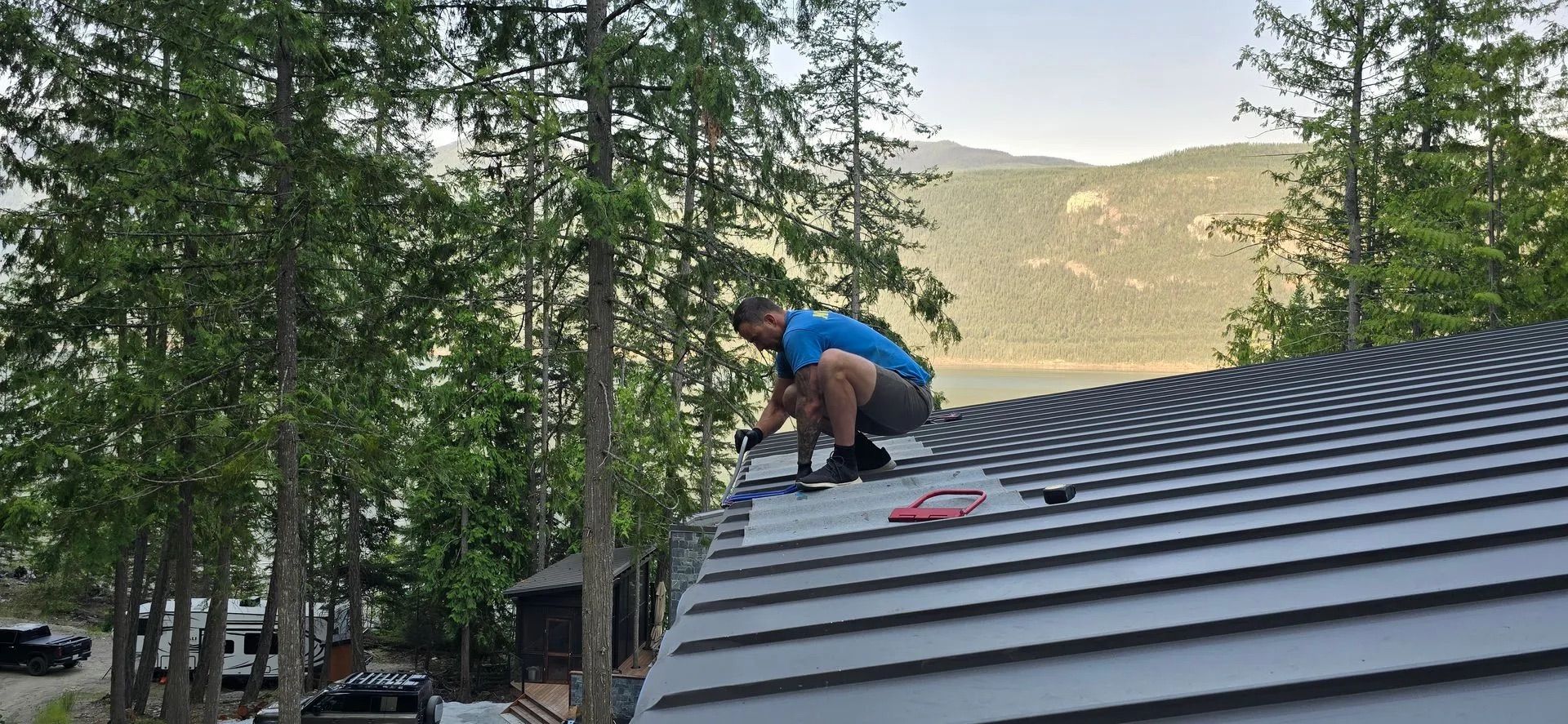 Worker crouches on a metal roof among pine trees with mountains in the background