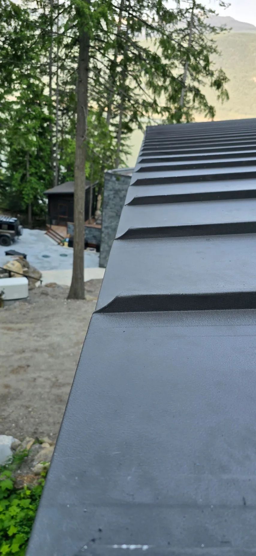 Steep black shingled roof beside trees and a house under construction