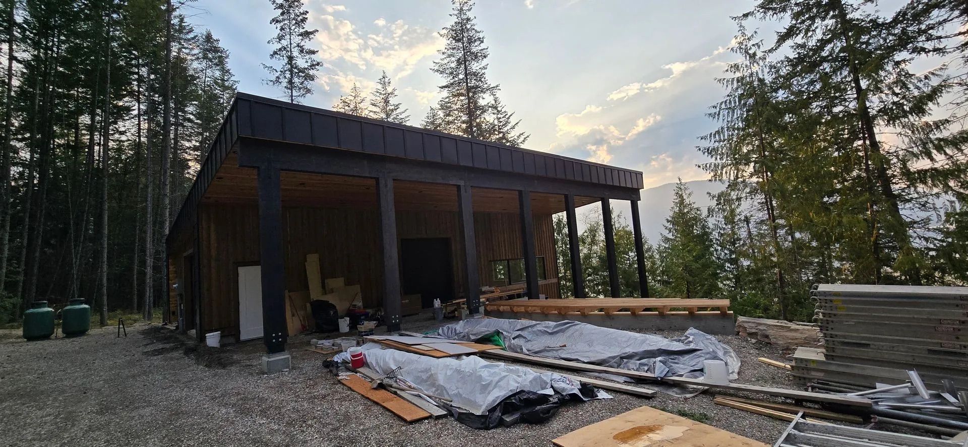 Partially built wooden cabin with open framing, tarp-covered ground, and tall trees at dusk