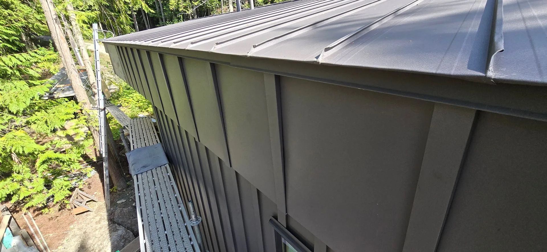 Side view of a gray metal roof and siding above a tree-lined yard.