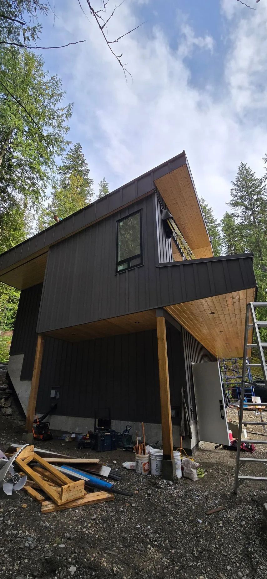 Modern black-and-wood cabin under construction in a wooded area, viewed from below under a cloudy sky