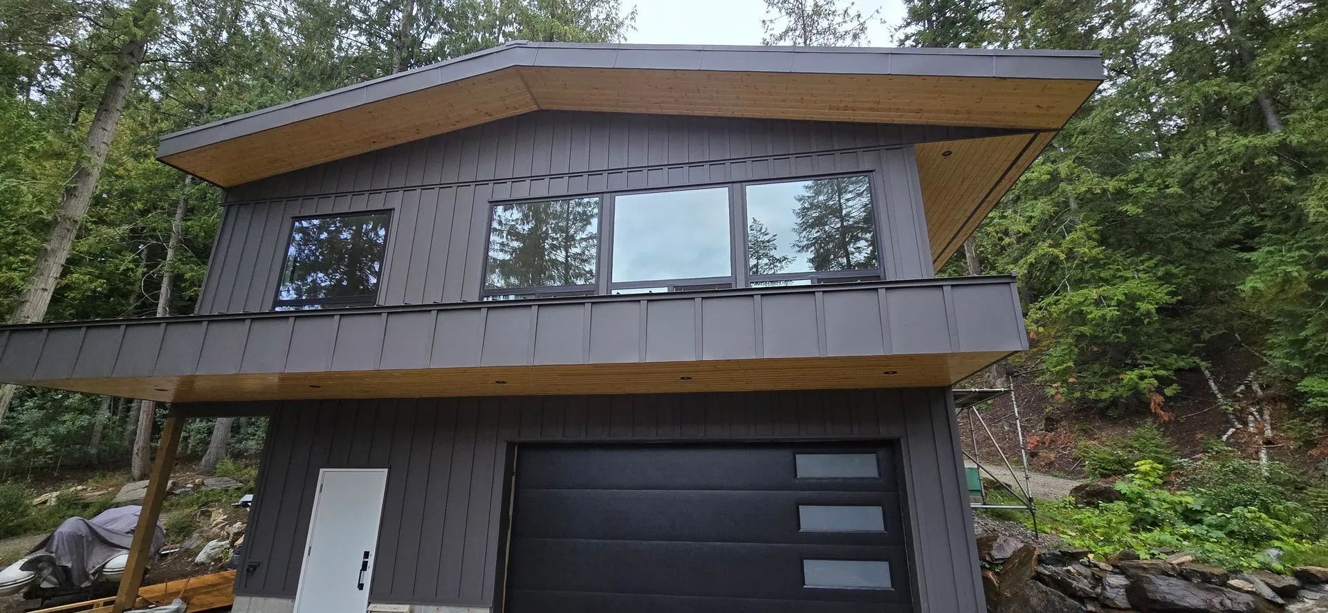 Modern two-story house with gray siding, large windows, and a black garage door amid trees