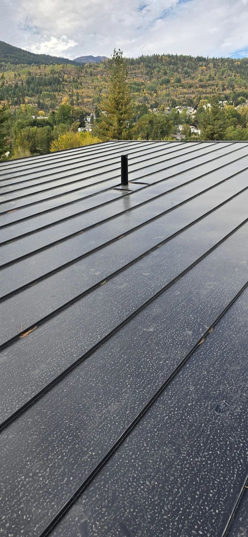 Dark metal roof with a vent pipe, overlooking green hills and cloudy sky