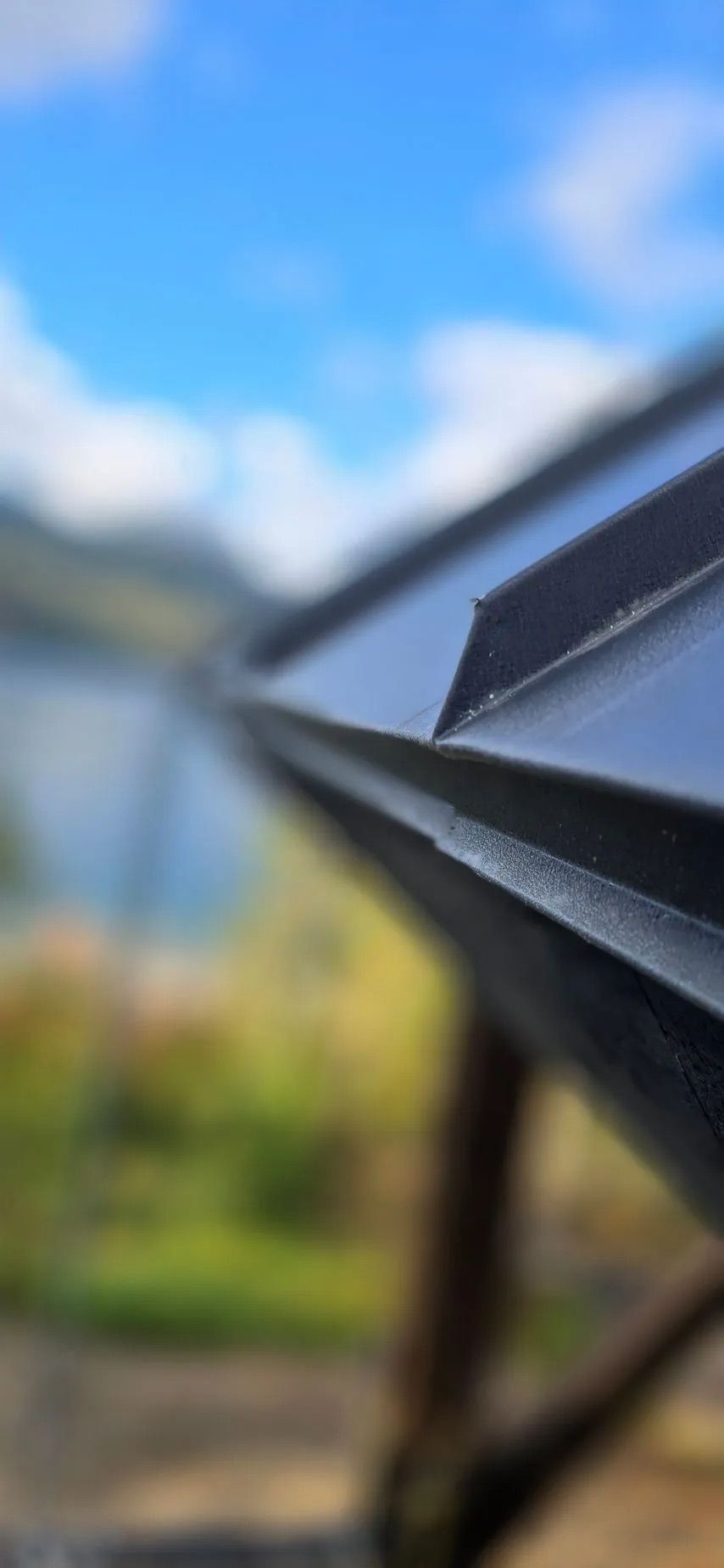 Close-up of black metal roof edge against a blurred blue sky and garden background
