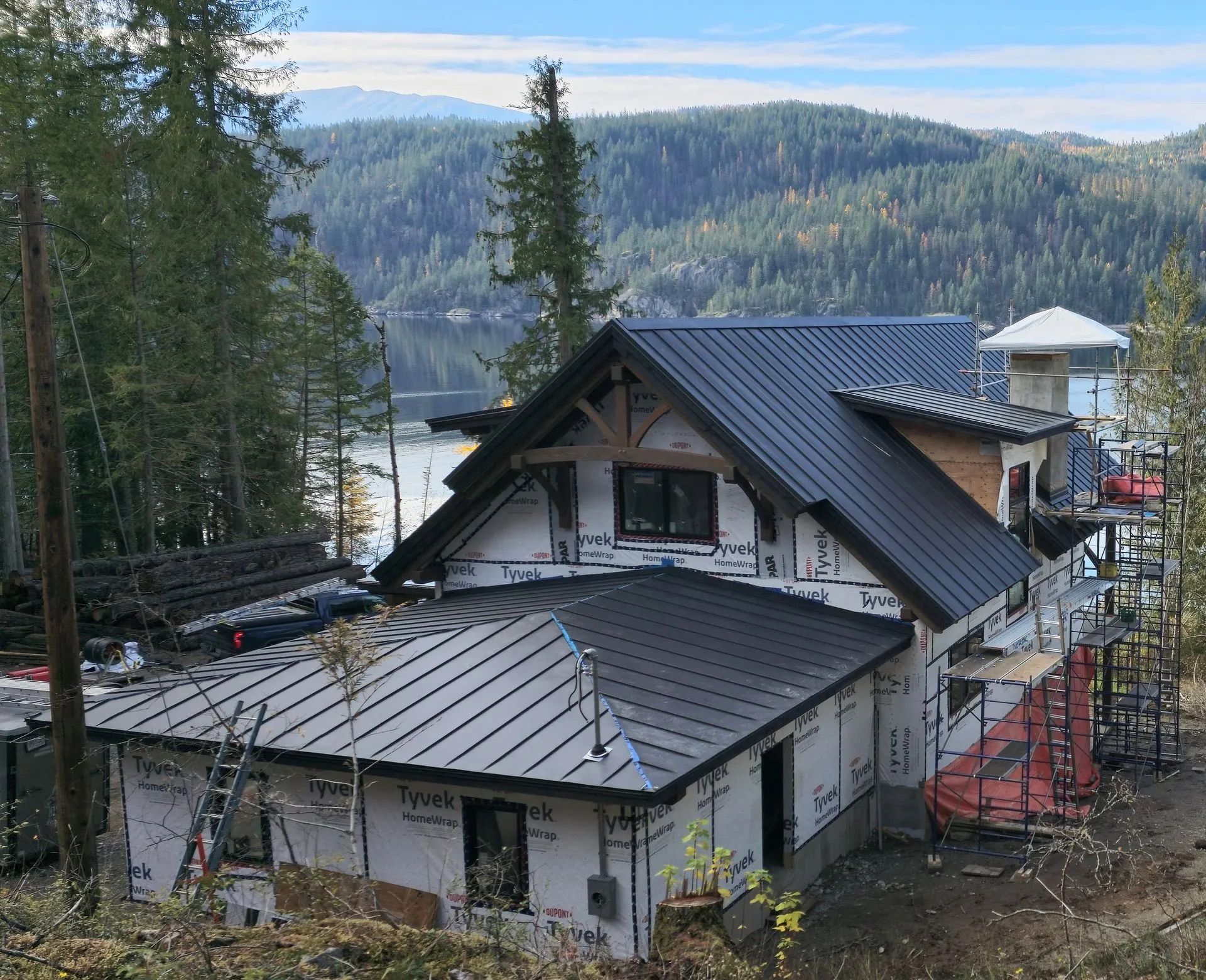 Mountain cabin with a dark roof on a forested hillside, overlooking a lake and distant pine-covered mountains.