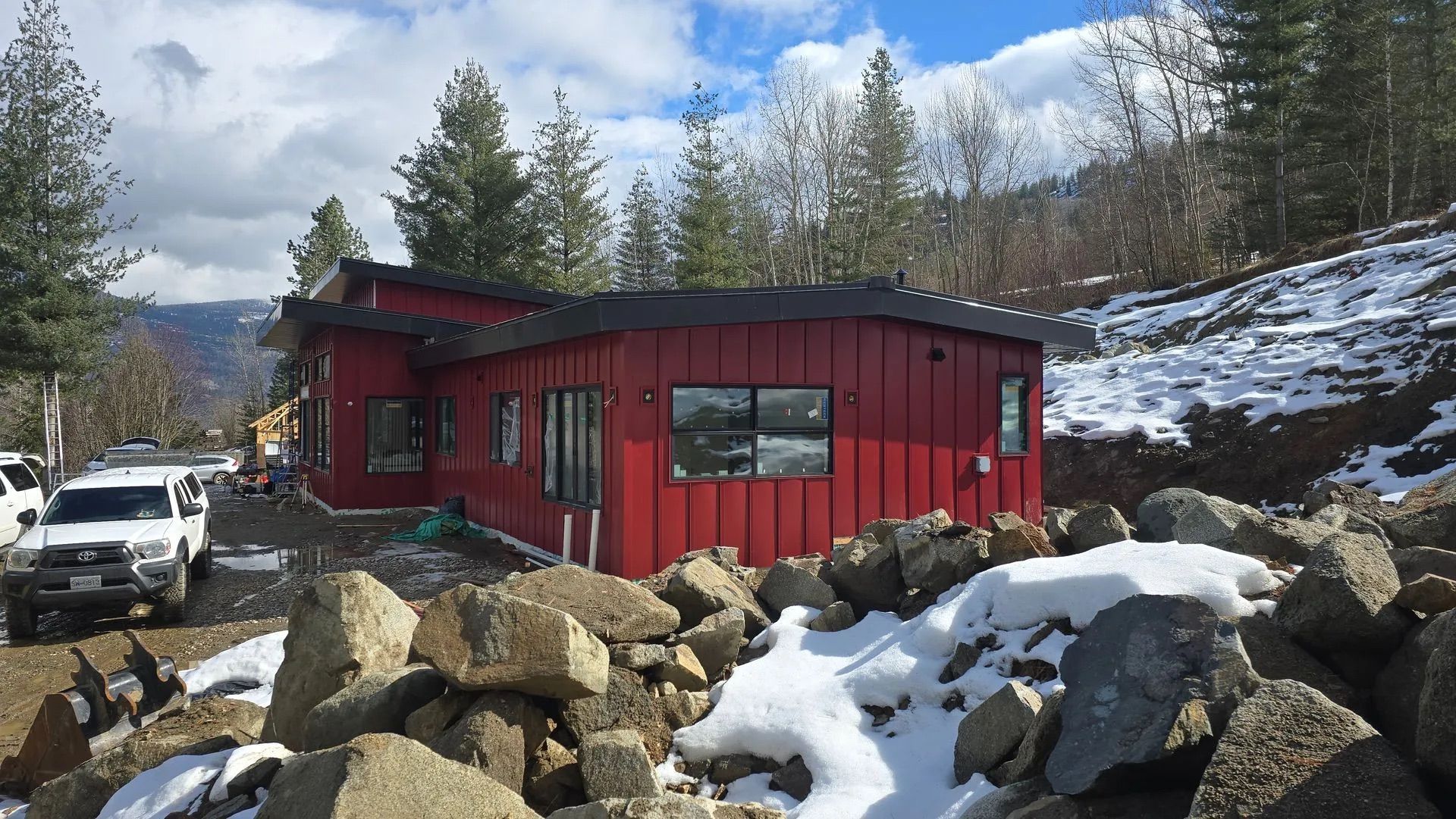 Red cabin in snowy rocky landscape with trees and parked cars under a cloudy sky