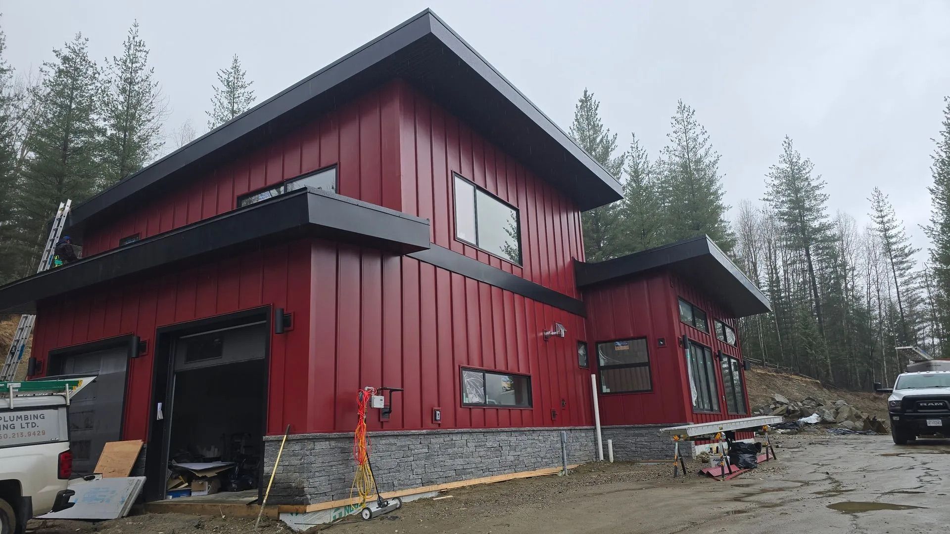 Red two-story house with black trim and stone base in a wooded, snowy setting