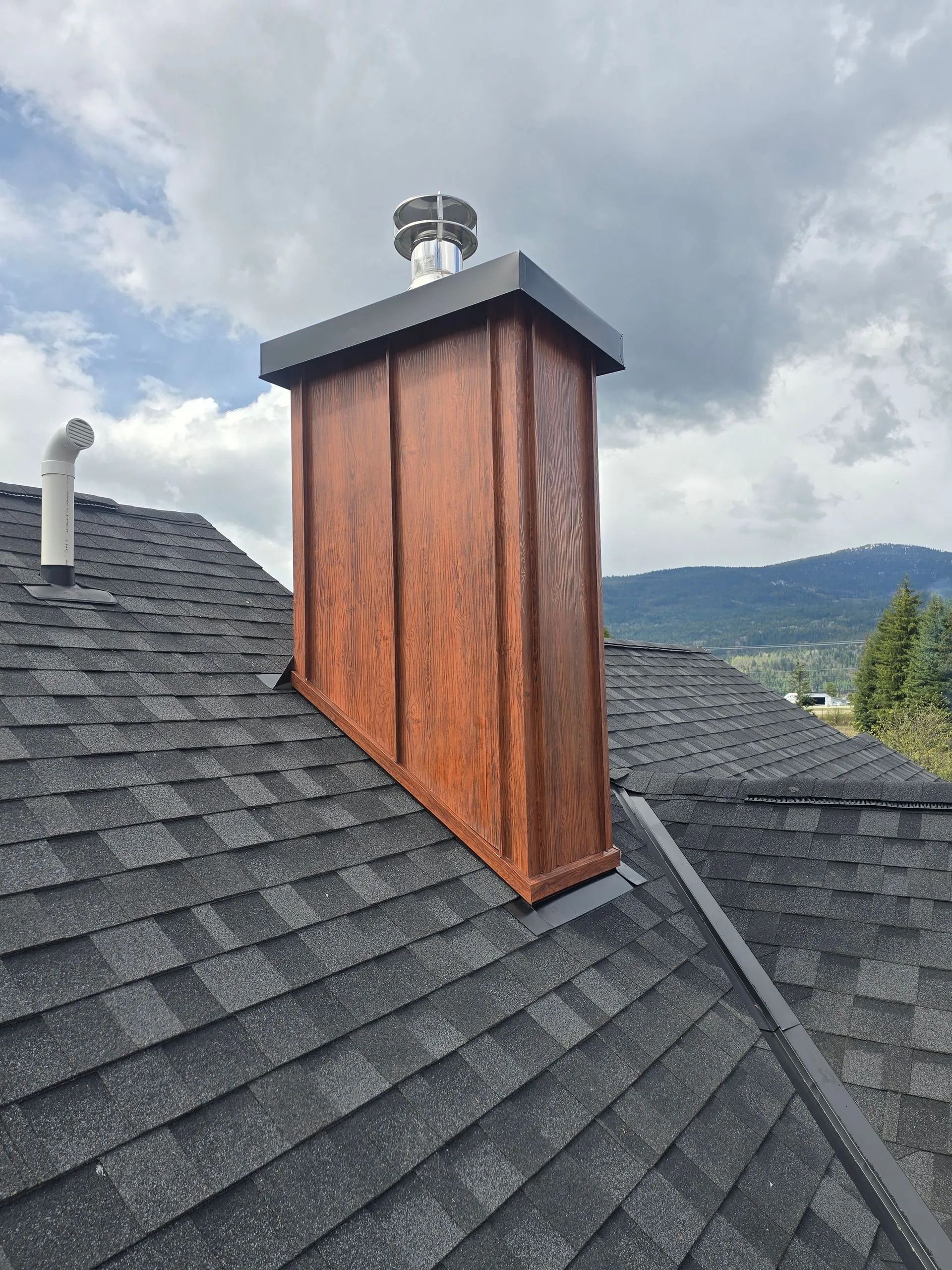 Roof with dark shingles and a copper chimney on a mountain home under a cloudy sky