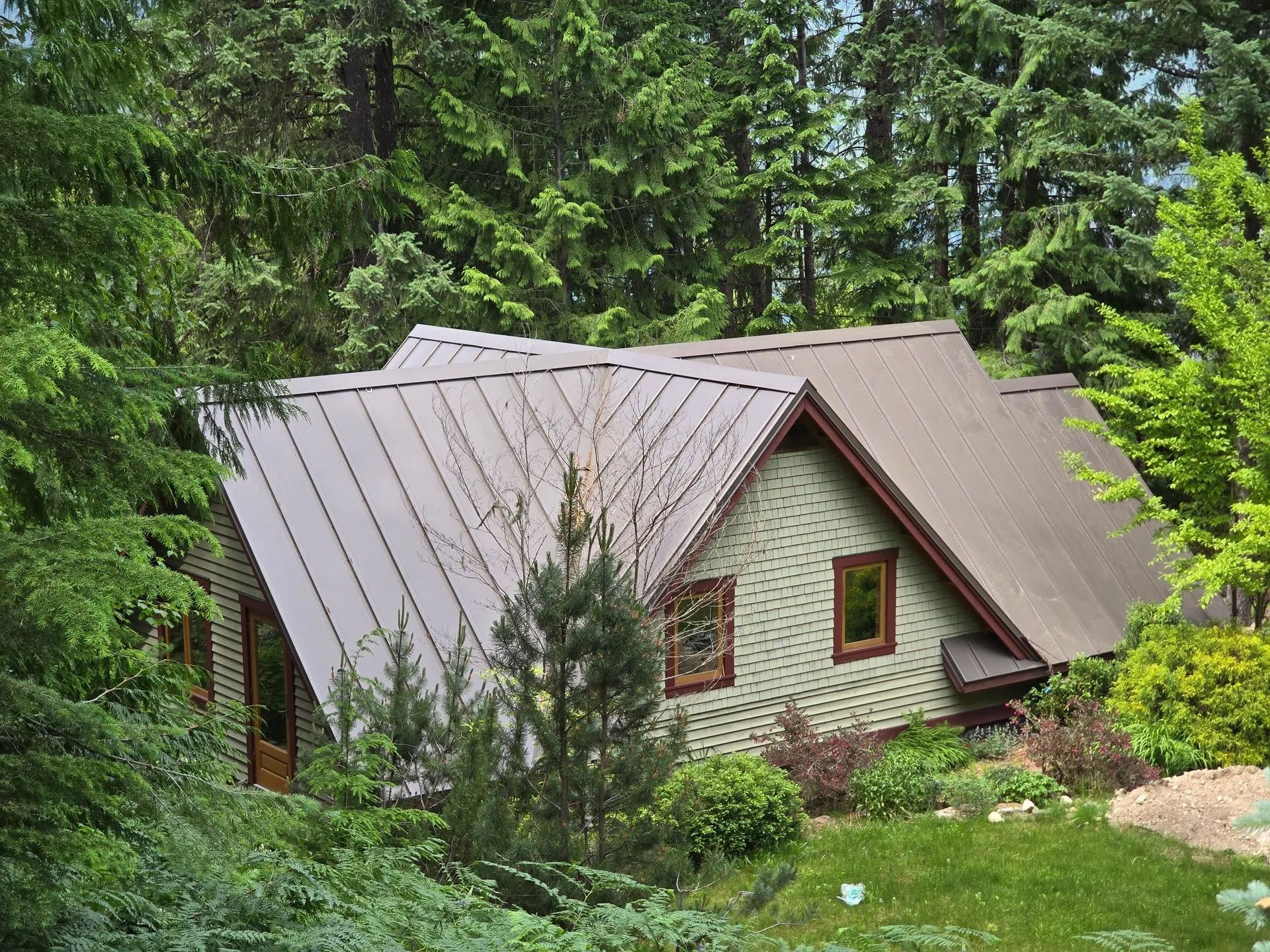 Small stone cottage with a gray metal roof nestled among dense green trees and shrubs.