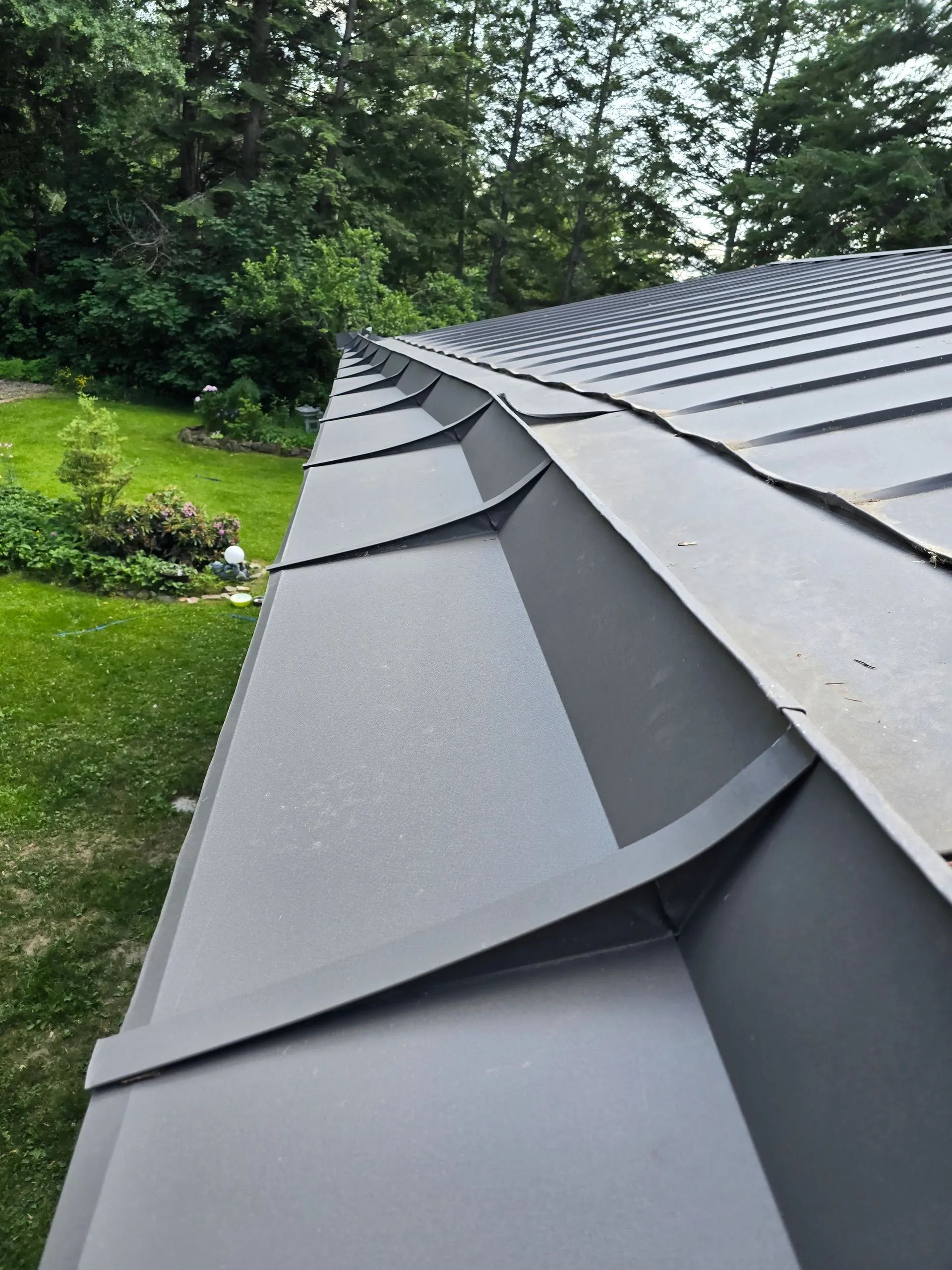 Close-up of a gray metal roof with standing seam panels and green trees in the background