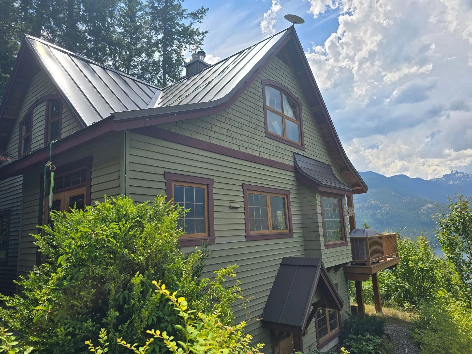 Green hillside cottage with metal roof, surrounded by trees, overlooking distant mountains under a cloudy sky