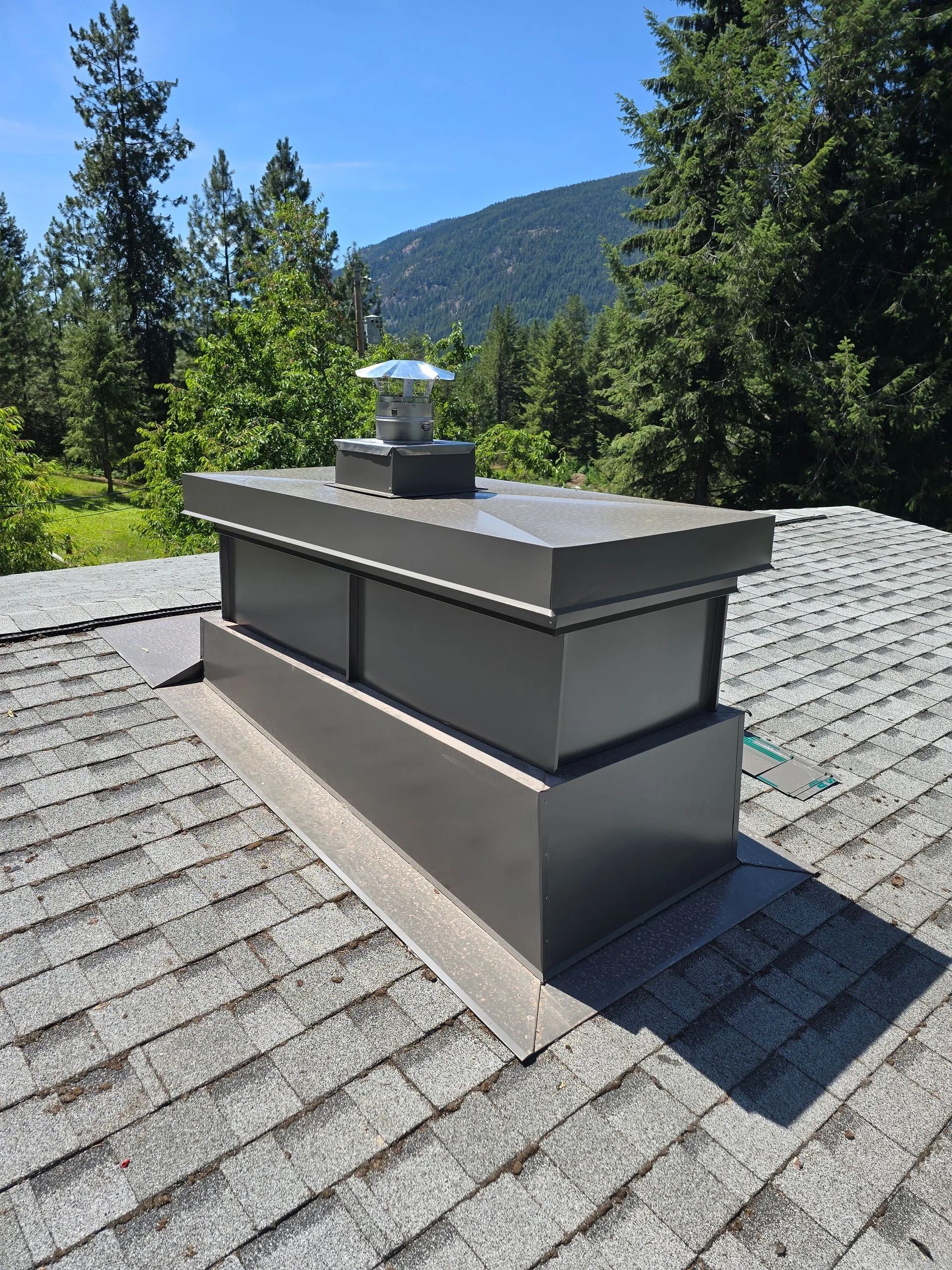 Rooftop chimney vent with gray cap on shingled roof, surrounded by pine trees and mountains.